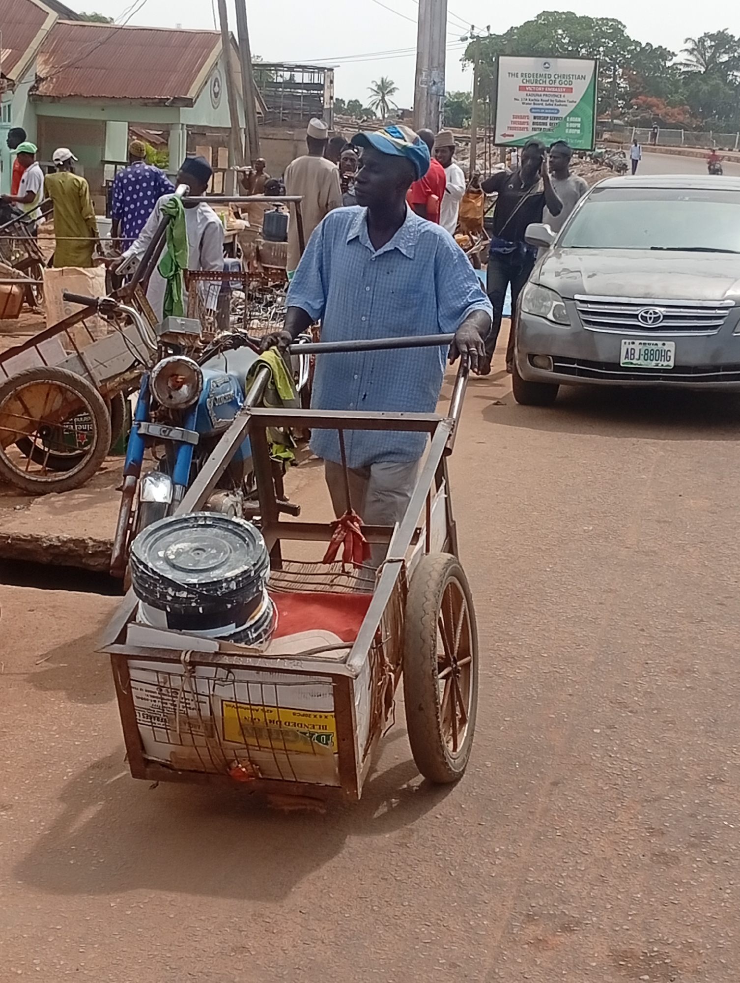 A man pushes a metal cart containing paint cans down a street in Sabon Tasha, Nigeria. The cart also carries what appears to be parts of a blue motorcycle. Various people are visible on the street, as is a gray car with license plate ABJ-880HG. A sign for The Redeemed Christian Church of God, Victory Embassy, is visible in the background. The environment appears to be an active commercial area.