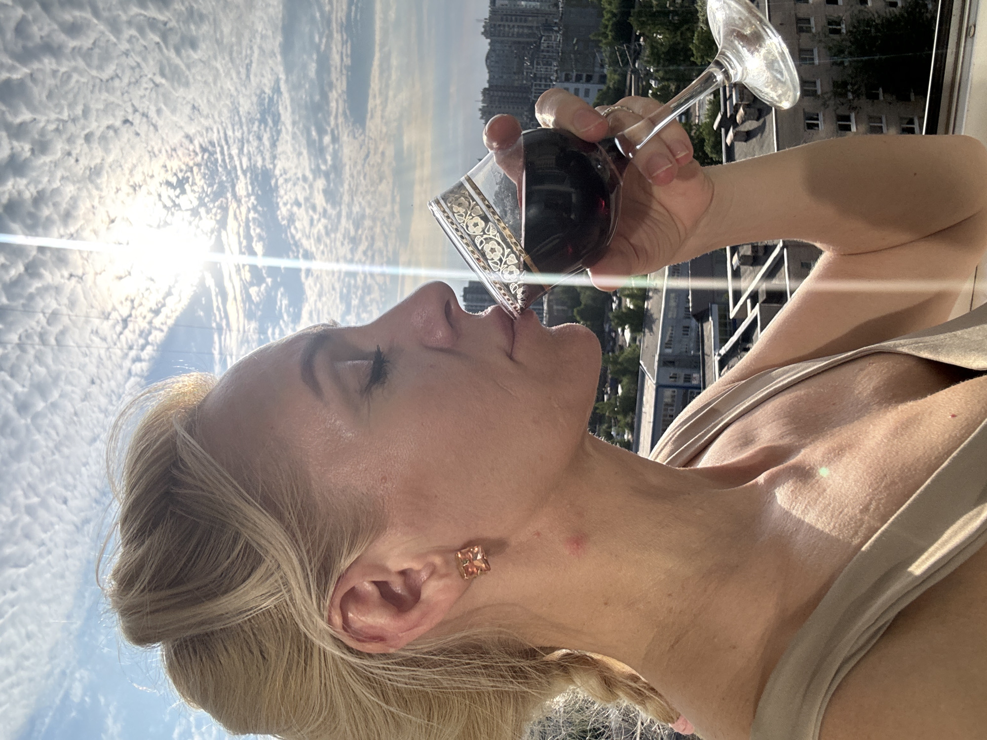 A blonde woman with her hair in a bun and a braid drinks red wine from a glass on a balcony overlooking a city in Russia. The sun shines brightly in a cloudy sky. She wears a tan sleeveless top and a square stud earring in her left ear. A red mark is visible on her neck. Buildings and trees are visible in the background.