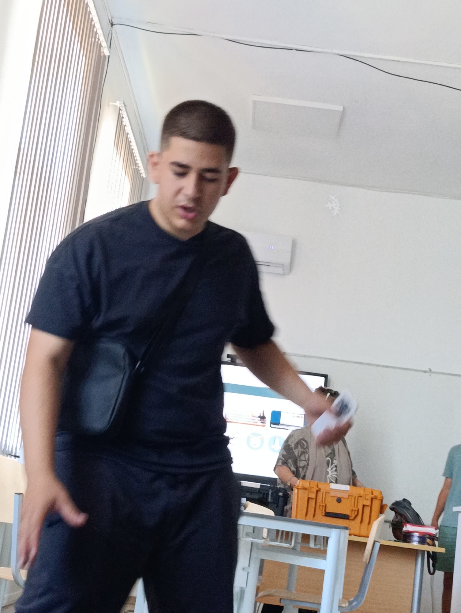 A young man wearing a dark t-shirt and dark pants, with a black cross-body bag, is in the foreground of an indoor scene. He is looking down and to his right, with his right hand holding a white object, possibly a phone or a card. Behind him, to the right, another person wearing a patterned garment stands near a desk with an orange case and other items. In the background, a projector screen displays some graphics, and an air conditioning unit is mounted on the wall. The environment appears to be a classroom or meeting room, indicated by the presence of desks, chairs, and educational materials. The lighting suggests it is daytime, with natural light coming from blinds on the left side of the frame.
