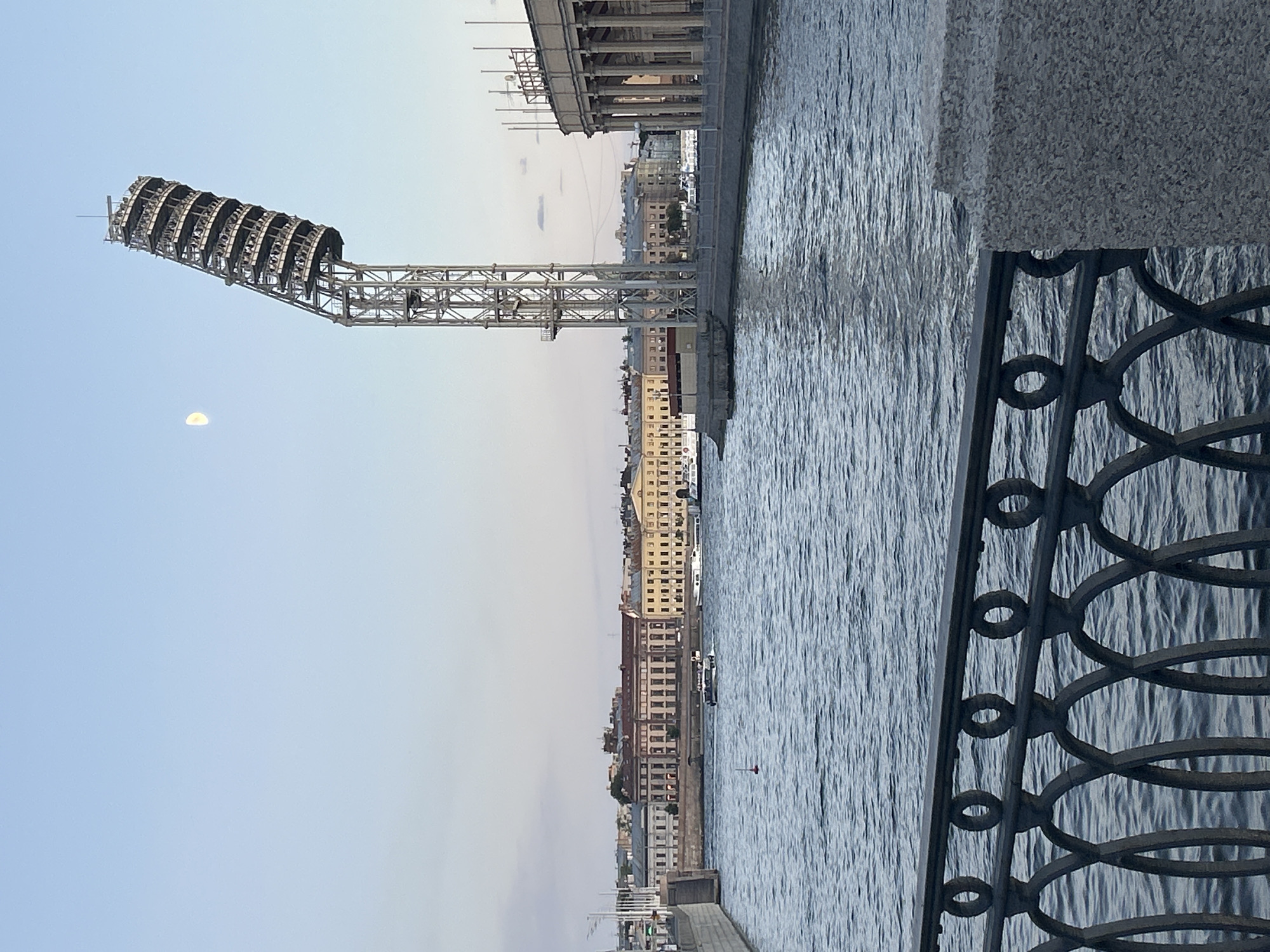 The image shows a view of a city waterfront with a large spotlight structure and a body of water. The spotlight, made of metal scaffolding, is positioned on the left side of the frame, extending upwards and outwards over the water. The moon is visible in the sky near the spotlight.
Across the water, a row of buildings with multiple windows lines the embankment. The architecture suggests an urban setting, possibly in Europe. The water is rippled, indicating some movement, and a dark, ornate metal railing runs along the right edge of the frame, separating the viewer from the water.
The sky is clear and light blue, with a gradient suggesting it's either early morning or late evening. There are no visible people, but the presence of buildings and a railing implies a public space. The overall mood is serene and contemplative, with the cool tones of the water and sky contributing to a sense of calm. No text is visible in the image.