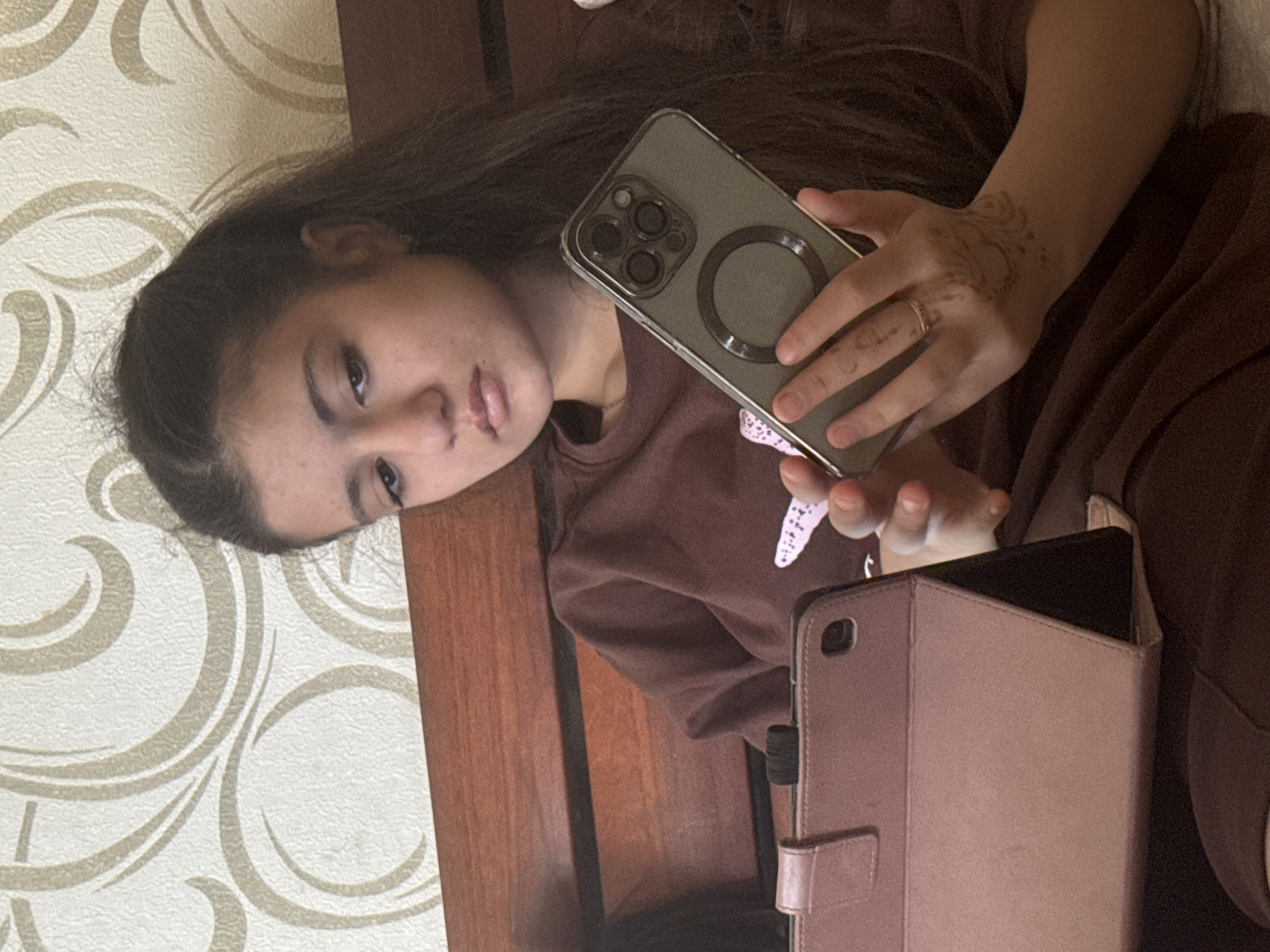 An adolescent or young adult female, with dark, long hair partially pulled back, is seated indoors. She wears a solid brown long-sleeved top and has a faint reddish-brown decorative design on the back of her right hand, along with a thin, gold-colored ring on her right ring finger. She holds a smartphone vertically in both hands, which is encased in a light olive/greyish-green protective cover featuring a prominent circular magnetic ring and a distinct camera module with three lenses and a flash/sensor array. Her gaze is directed downwards towards the phone. To her left, a tablet in a light pink/rose gold foldable case is propped up in landscape orientation, suggesting passive viewing or use. The background consists of light-colored (off-white/cream) wallpaper with a repeating curvilinear, ornamental pattern in a beige/light brown color, situated above a horizontal wooden structure with visible grain. The scene is located in Unknown, Unknown.
