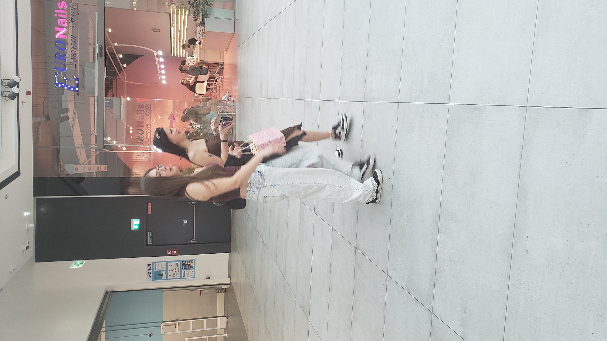 The image captures a moment within an indoor shopping mall, likely in Prešov, Slovakia, based on the presence of the "ACTION" store, a common chain in Central Europe. Two young women are walking side-by-side, their figures slightly blurred due to motion. The woman on the left, with long brown hair, wears a dark halter top and loose-fitting, light-wash jeans. She carries a dark bag over her left shoulder and is looking forward. The woman on the right, also with long brown hair, sports a strapless brown top and a dark skirt, carrying a pink and gold patterned shopping bag in her right hand. She appears to be looking to her left.
Behind them, a storefront for "EURO Nails American Style" is visible, featuring neon signage and illuminated displays. Inside the nail salon, several customers are seated at manicure stations, being attended to by staff. The mall's flooring is tiled with large, light-colored squares. To the left, a section of the "ACTION" store is visible, with signage and a shelf unit displaying products. Emergency exit signs with green arrows are present on the wall, indicating egress routes. The overall environment suggests a well-lit, modern commercial space.