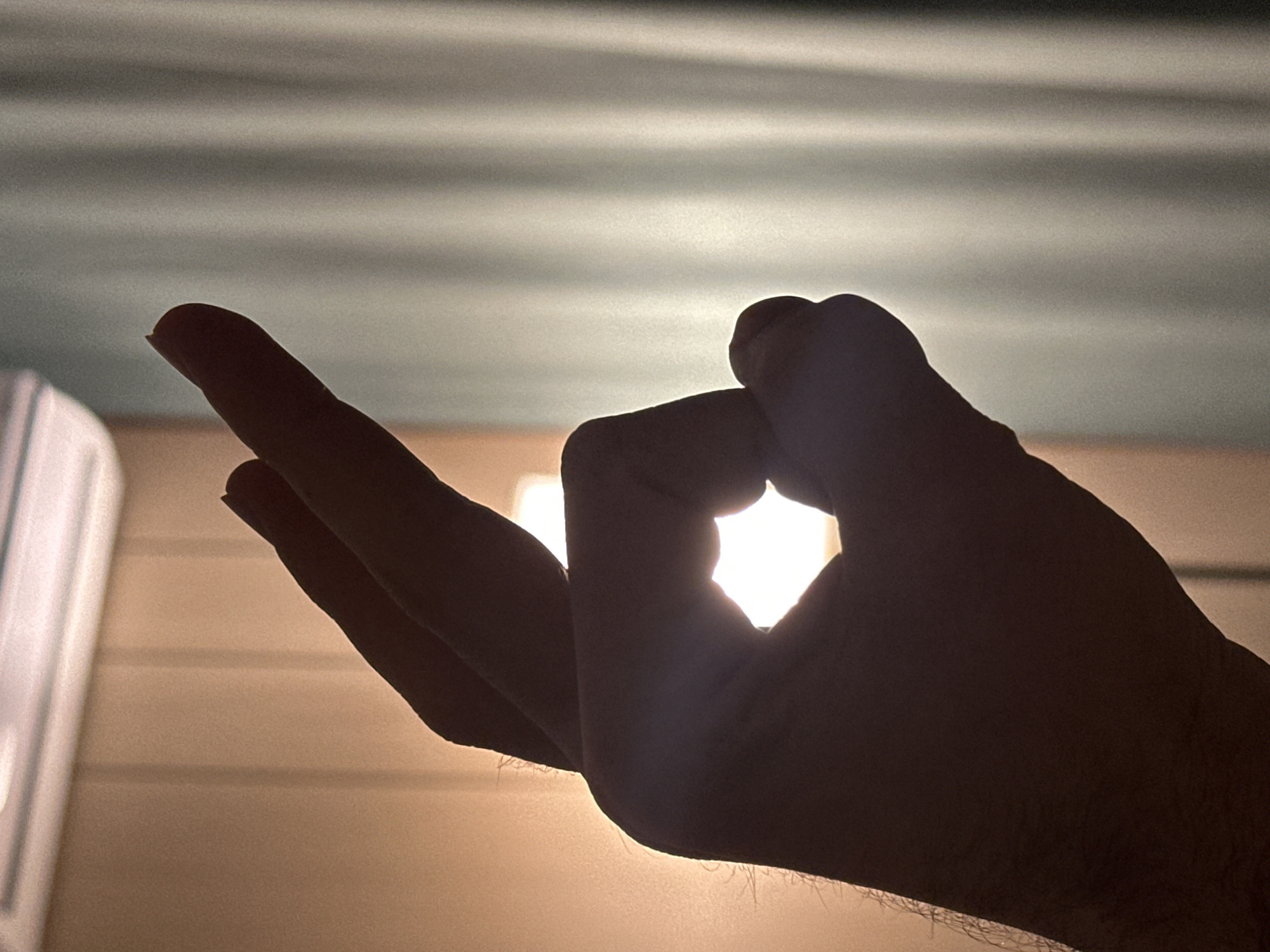 A close-up shot shows a silhouetted hand with the index finger and thumb forming a circle, backlit by a bright light source, creating a halo effect. The middle and ring fingers are partially visible. The background consists of an interior space with horizontal lines, suggestive of a wall or ceiling. The location is unknown and not suggested in the image.