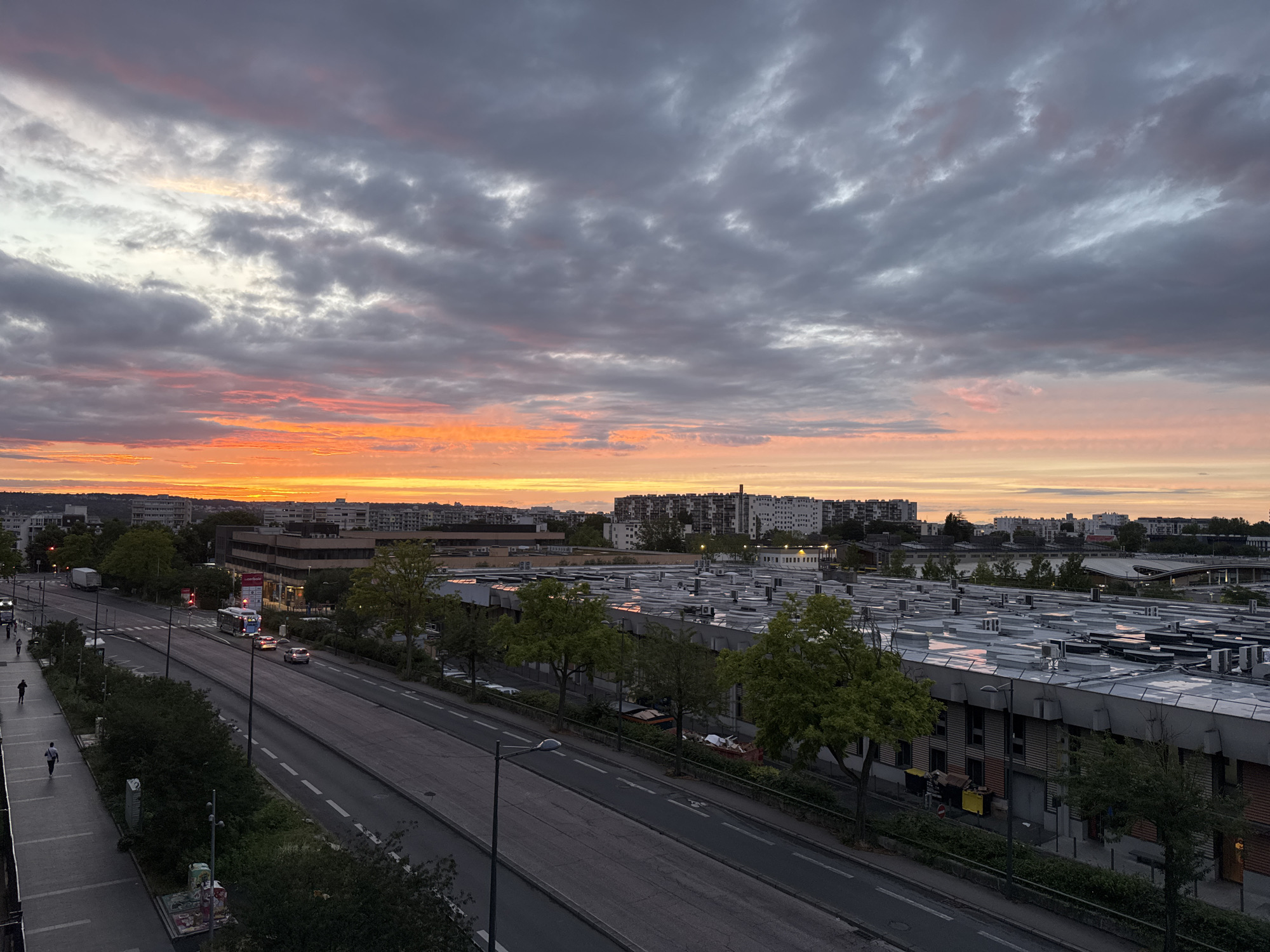 This is an aerial, wide-angle photograph capturing a sunset over the city of Fresnes, France. The sky dominates the upper portion of the image, displaying a dramatic gradient of colors from deep grey clouds at the top to bright orange and yellow hues near the horizon. A layered expanse of clouds, tinged with pink and orange, stretches across the sky.
Below the sky, a cityscape unfolds. In the foreground, a multi-lane road runs diagonally from the lower left to the upper right. The road has white lane markings and a streetlamp on the right side. A sidewalk is visible on the left of the road, with two individuals walking, one in a dark shirt and the other in a light shirt. Further down the sidewalk, two more individuals are walking. A few trees with green foliage line the sidewalk.
Across the road, a row of buildings is visible. The most prominent structure is a long, low-rise building with a light-colored, corrugated roof, dotted with numerous air conditioning units or vents. Several trees with green foliage are situated in front of this building. Beyond this building, a cluster of taller, residential buildings with white facades can be seen in the mid-ground. In the distance, more apartment buildings are visible against the horizon.
Traffic on the road includes a bus, a truck, and several cars. Red traffic lights are illuminated at an intersection. In the lower right portion of the image, a dumpster filled with debris is visible next to a building. The overall scene depicts an urban environment during sunset, with a mix of residential and industrial or commercial structures.
