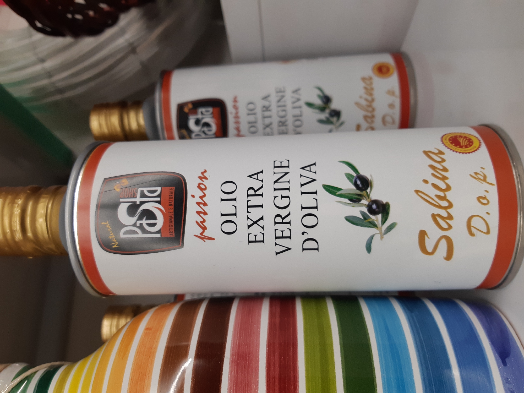 The scene depicts three containers of liquid food products arranged on a white shelf. The foreground is dominated by a white, cylindrical metal tin of "Sabina Olio Extra Vergine D'Oliva" with a gold screw cap. Its label features "Natural PaSta ARTIGIANALE E NATURALE" at the top, "passion" in red script, "OLIO EXTRA VERGINE D'OLIVA" in black text, an illustration of an olive branch with two black olives and green leaves, and "Sabina D.o.p." with a circular gold seal at the bottom. Red bands mark the top and bottom edges of the label and the container. To the immediate left, a portion of a ceramic bottle or jar is visible, characterized by prominent horizontal stripes in various colors including brown, red, green, blue, and purple, also topped with a gold cap. In the blurred midground to the right, a second identical "Sabina Olio Extra Vergine D'Oliva" tin is visible. The immediate environment consists of the white shelf, with a plain light-colored background wall or additional shelving units. A dark, textured object is partially visible in the upper right background. The location is an indoor retail or pantry setting within Plovdiv, Bulgaria. No individuals are present in the scene.