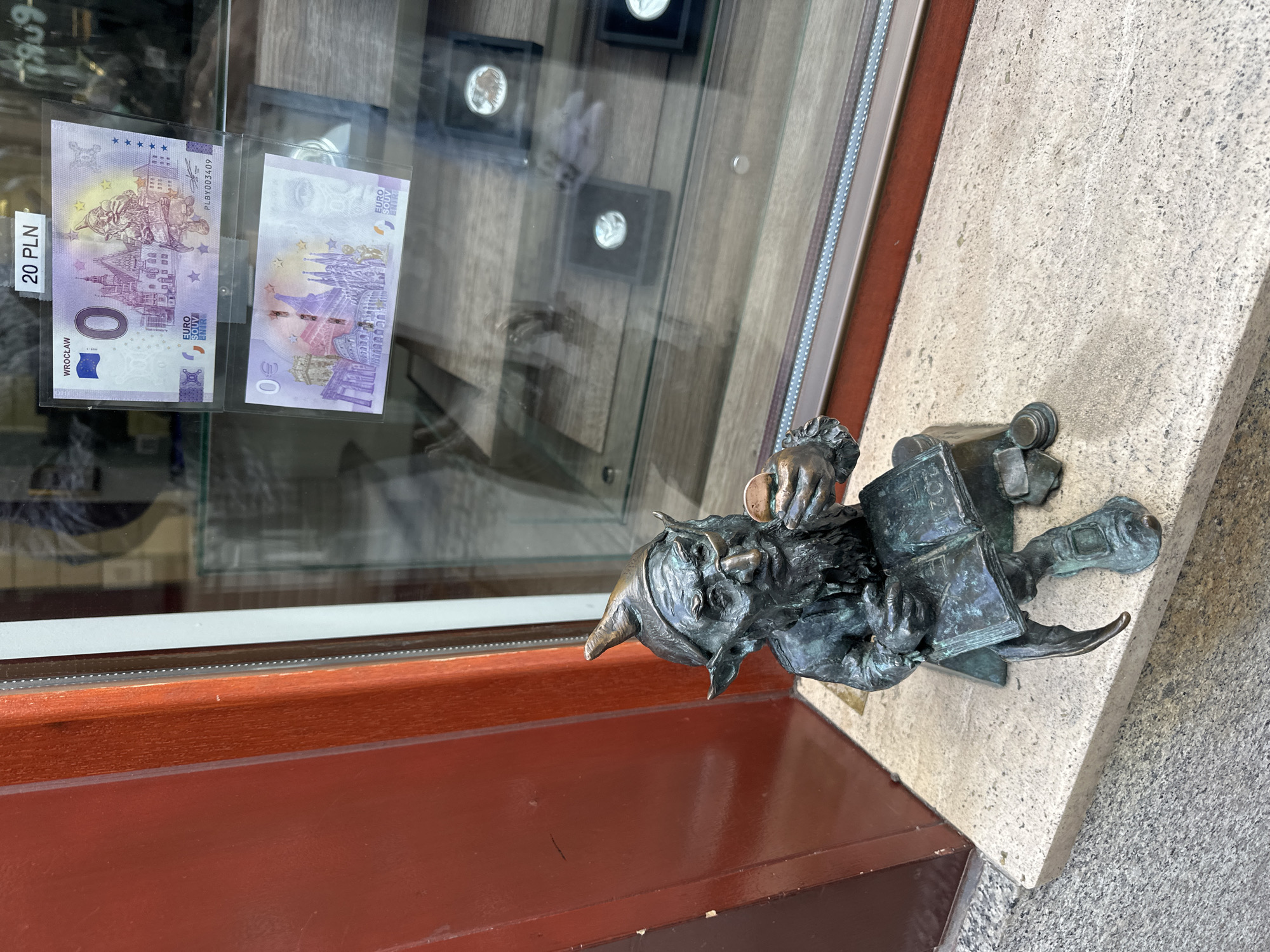 Here is a description of the image:
The image shows a small bronze statue of a gnome or dwarf, situated next to what appears to be a shop window or display. The gnome is holding a book and is positioned on a light-colored, weathered stone surface. In the background, behind a glass panel, are what seem to be souvenir banknotes, specifically a "0 Euro" note with "Wrocław" printed on it and labeled "20 PLN". There are other framed items, possibly coins, displayed in the background as well. The setting seems to be outdoors, with the statue placed on a ledge or windowsill next to a building. The lighting is natural, and the scene is captured at eye level, suggesting it's a ground-level display or exhibit in the city of Wrocław, Poland.