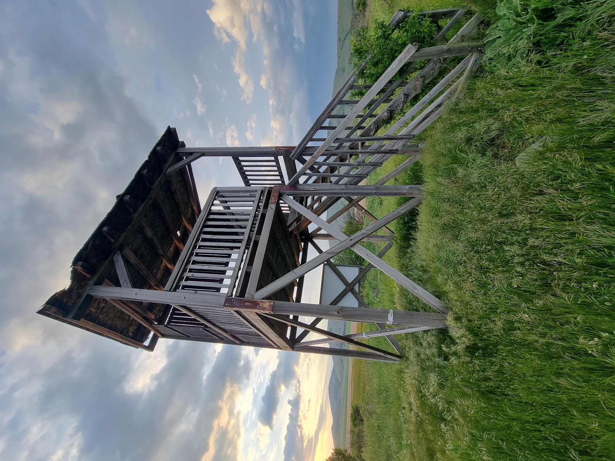 The image depicts a weathered, wooden watchtower or observation platform, likely abandoned, set in a grassy field near Aldomirovtsi, Bulgaria. The structure has two levels with a partially dilapidated roof and stairs leading to the upper level. The sky behind it features a mix of clouds and sunlight, suggesting either sunrise or sunset. The surrounding environment consists of tall grass and some vegetation. In the background, a low hill can be seen. No people are visible in the image.