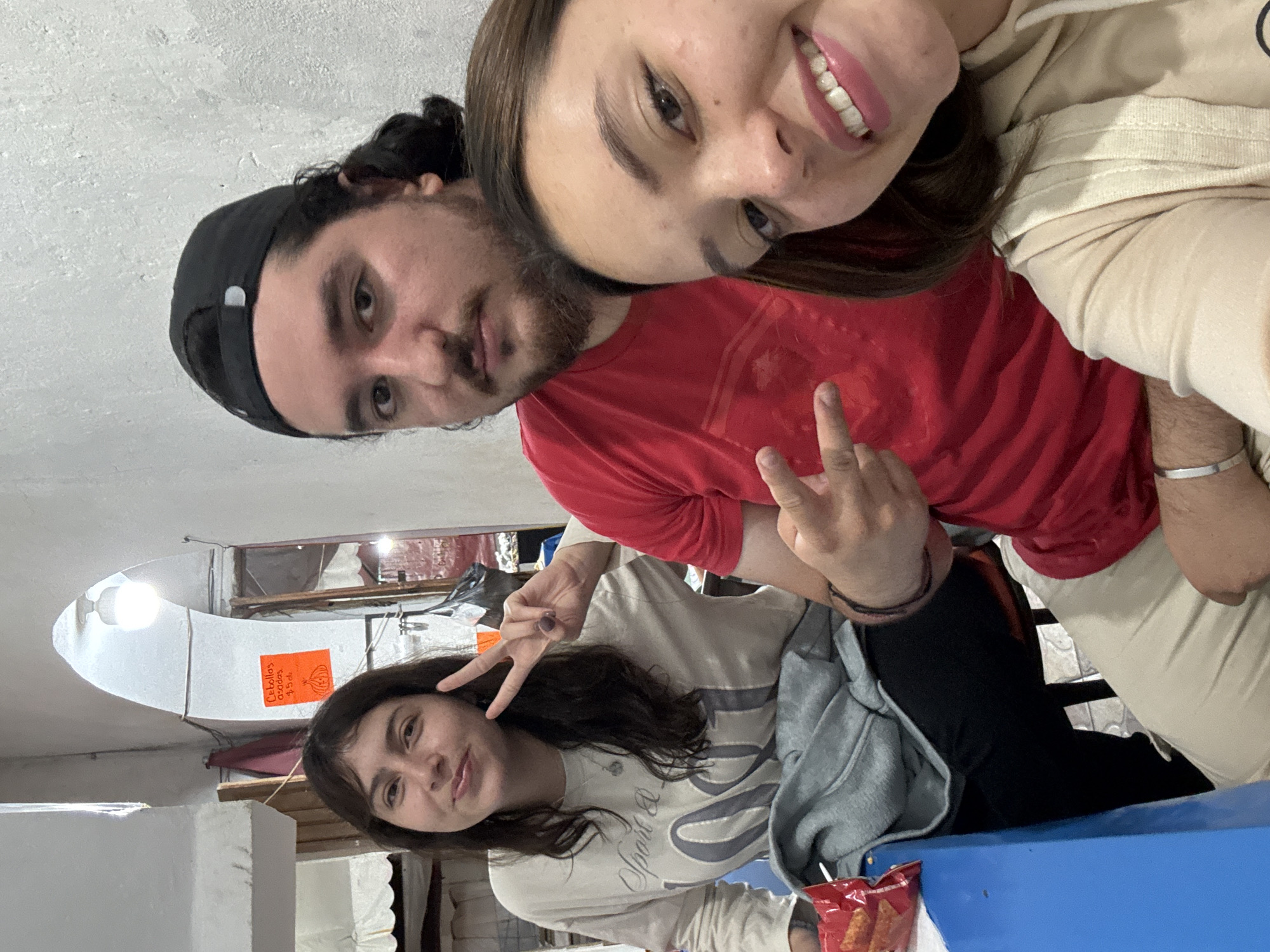 Three individuals are captured in a selfie taken indoors. The image appears to be rotated counter-clockwise. At the top, a light-skinned male with a beard and a black baseball cap is wearing a red shirt and making a peace sign. He is positioned behind a fair-skinned female, who is smiling. The woman is also wearing a tan hooded sweatshirt.
Below, there is another woman with dark hair and dark eyes who is also making a peace sign. She is wearing a light colored shirt. There are also chips on the table next to her.
In the background, there is what looks to be a market, with an overhead fluorescent lighting fixture. A sign is written in Spanish and the market is inside. The location is likely the city of Cuetzalan, Mexico, as indicated in the instructions.