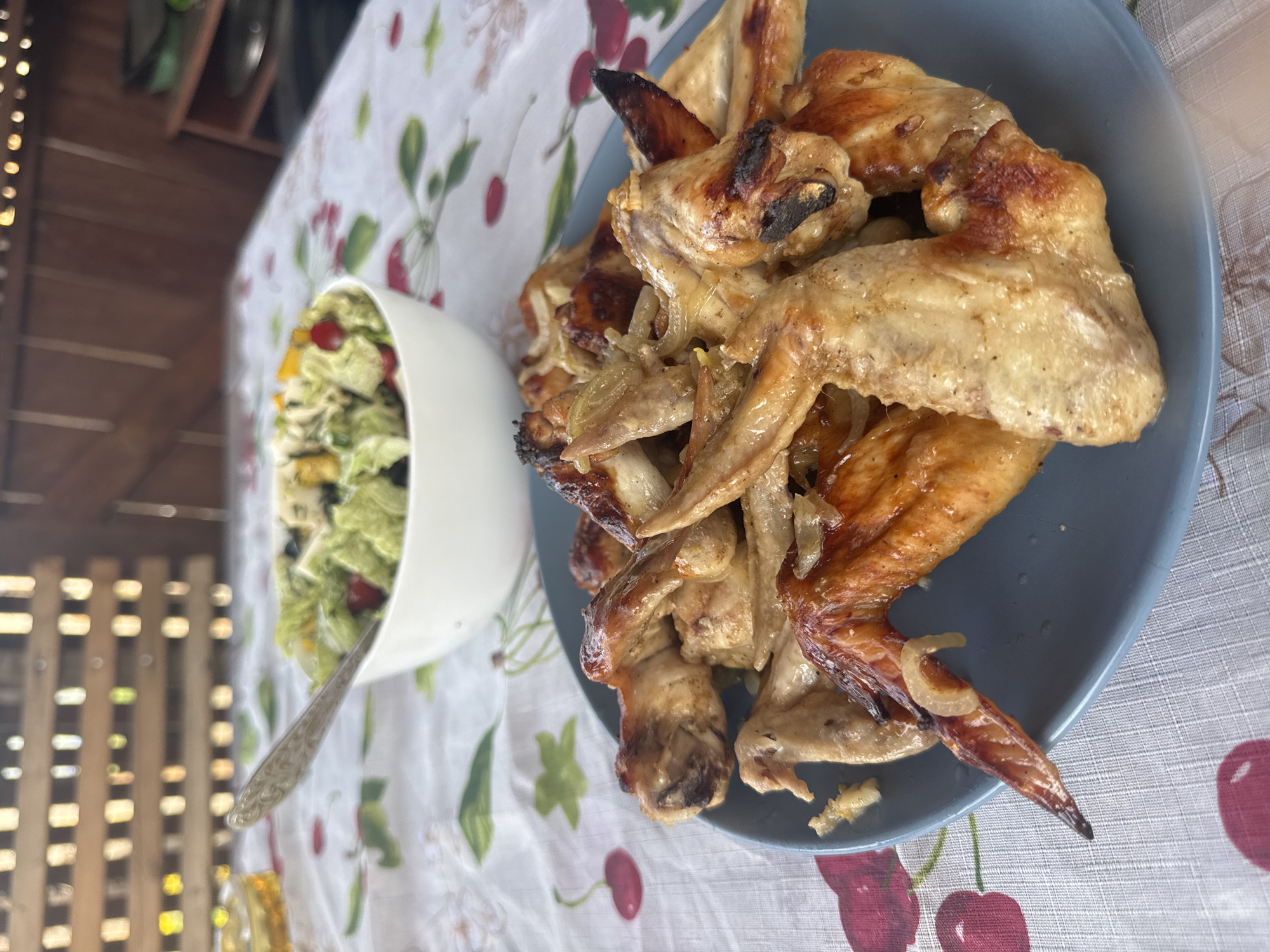The image shows a plate of cooked chicken wings and a bowl of salad on a table covered with a patterned tablecloth. The scene is outdoors, with a slatted structure visible in the background. The precise location is not discernible from the image, but a city in Russia, Chaikovsky, is assumed.