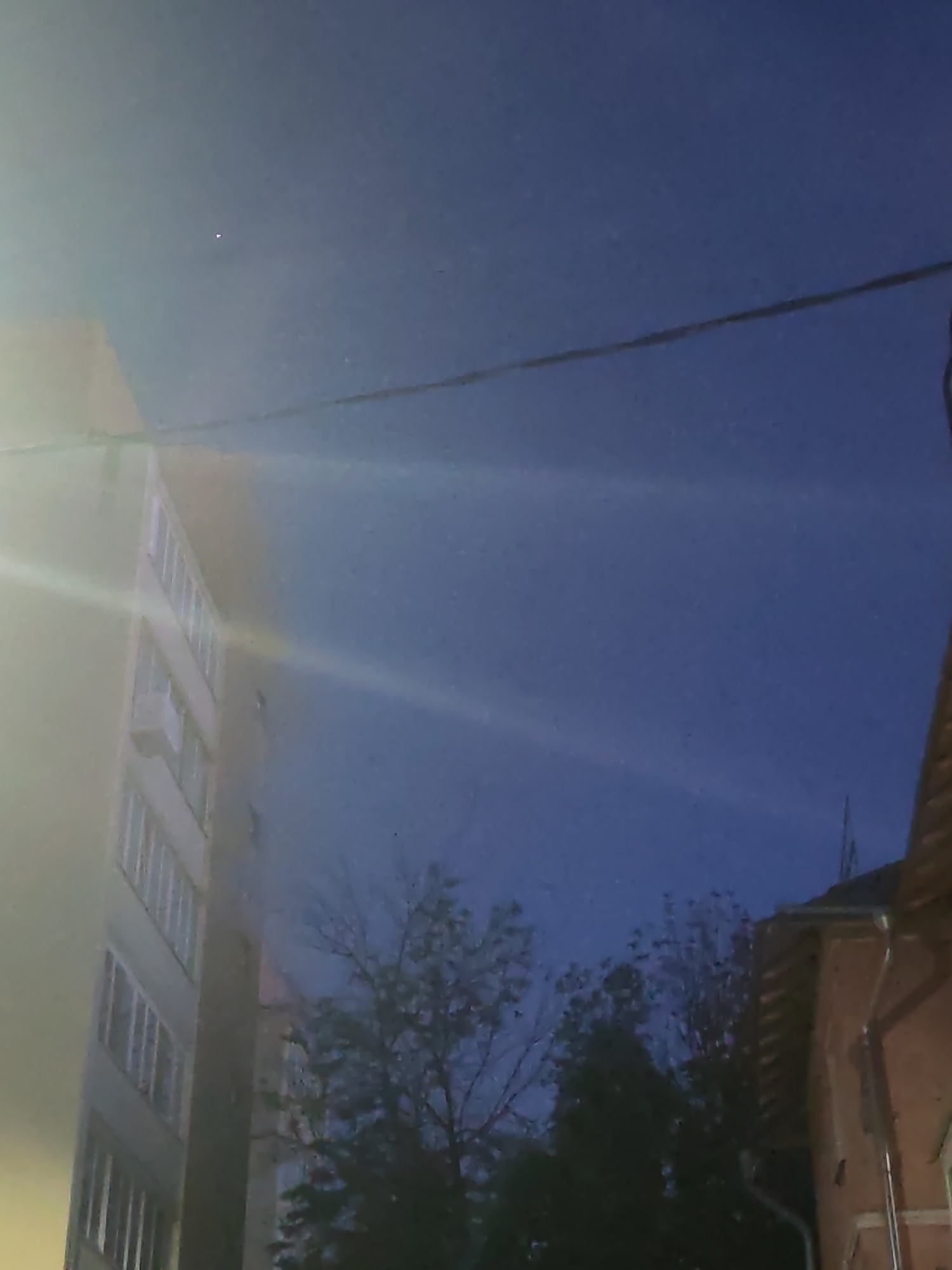 The image shows a twilight sky with a gradient from a brighter, yellowish hue near the left edge to a deep blue across the majority of the frame. A thin, dark power line stretches horizontally across the upper portion of the image. On the left, a portion of a modern apartment building is visible, characterized by its light-colored facade and repeating rows of windows. A strong light source, possibly a street light or a reflection, creates a lens flare effect across the building and into the sky. In the lower right quadrant, a section of a brick building with visible gutters is present. Between the buildings, bare-branched trees are silhouetted against the darkening sky. The scene appears to be an urban setting at dusk or early night.