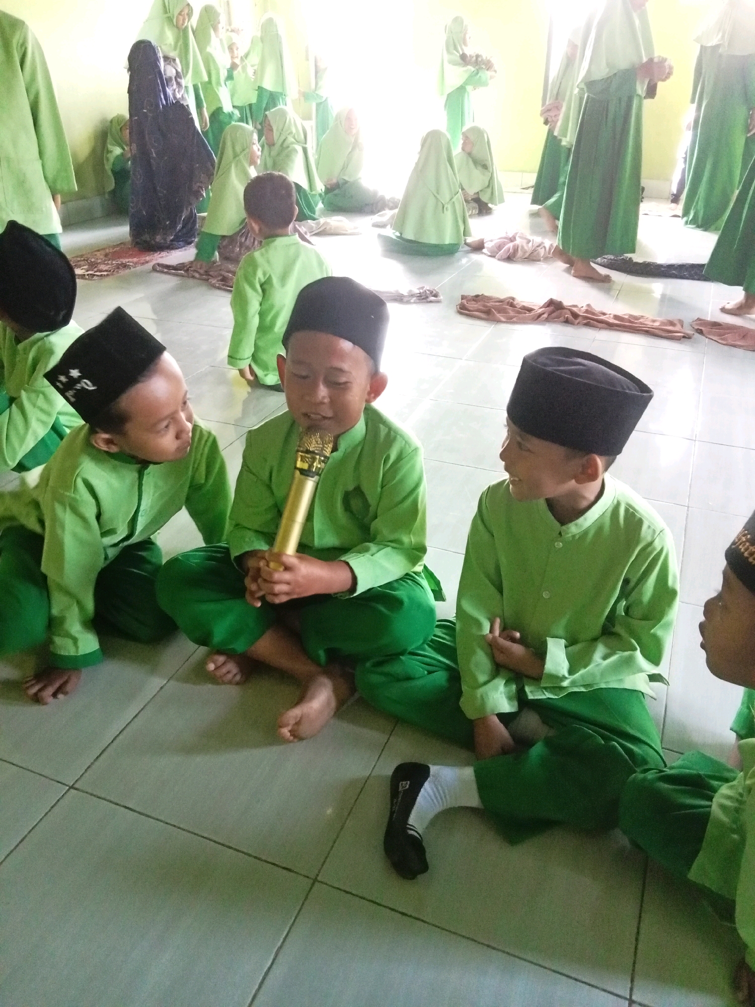 In Lebo Jaya, Indonesia, a group of children, dressed in traditional Islamic attire of green long-sleeved shirts and pants, and black peci hats, are gathered in what appears to be an indoor space with tiled flooring. Three boys are prominently positioned in the foreground, seated on the floor. One boy, holding a gold microphone, is actively speaking or singing. Another boy next to him is looking at the speaker, his head tilted. The third boy, to the right, is looking away from the speaker and smiling. In the background, several other children and adults, also in similar Islamic clothing, are visible, some standing and some sitting. The lighting suggests an indoor environment, possibly a school or community hall, with natural light coming from the left. The overall scene depicts a religious or cultural gathering with a focus on a performance or recitation by the children.