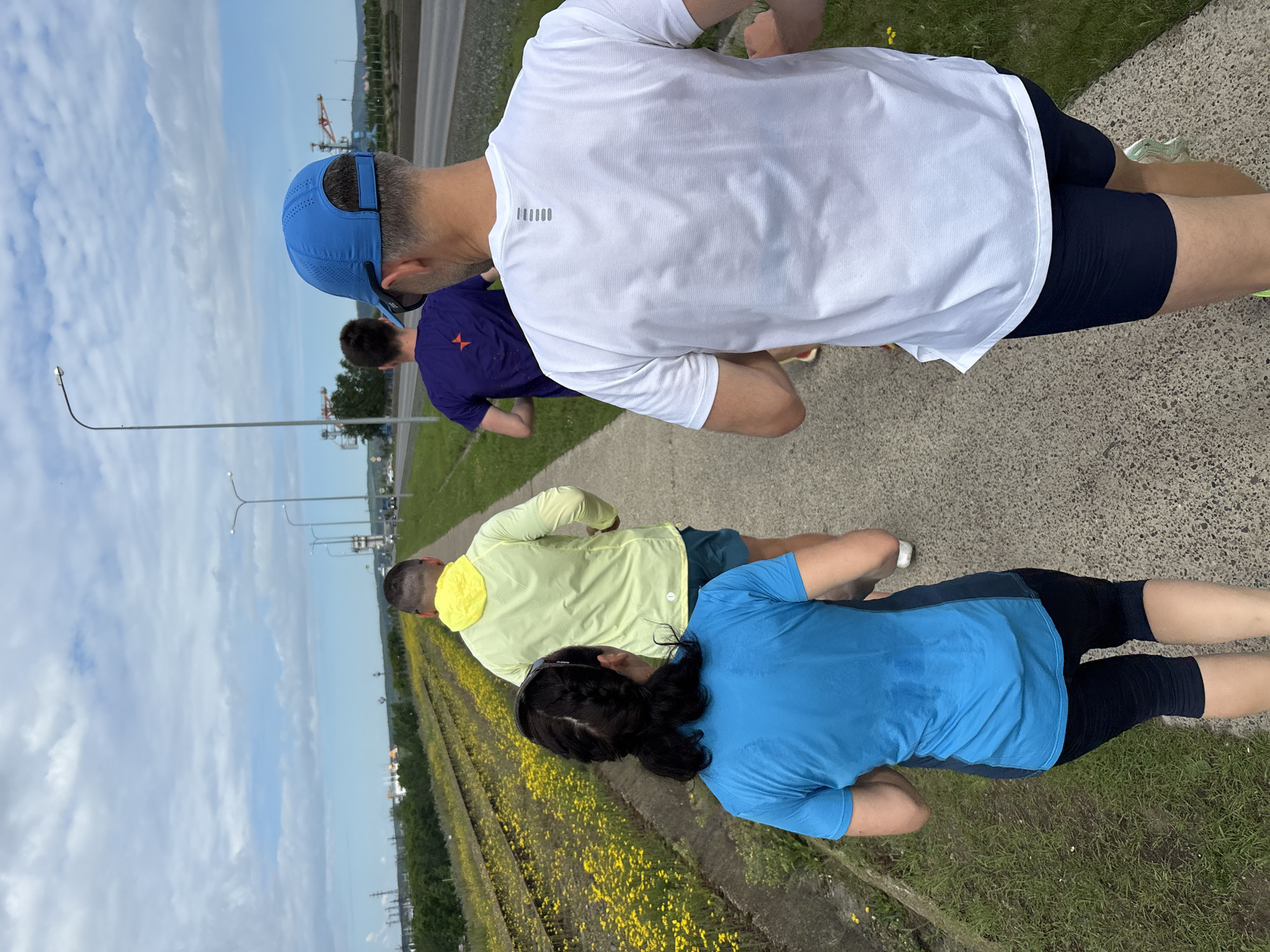 Four individuals are depicted running on a paved pathway in Chaikovsky, Russia, viewed from behind as they move away from the camera. The foreground shows the backs of four runners: a female in a bright blue short-sleeved athletic top exhibiting sweat marks on the back, with her dark hair braided, and wearing dark cropped pants; a male in a bright yellow long-sleeved hooded athletic top and dark shorts; a male in a dark purple short-sleeved athletic shirt featuring a red star-like emblem on the upper back, and dark shorts; and a male in a white short-sleeved athletic shirt with a subtle grey vertical logo on the upper back, wearing a blue perforated baseball cap backwards, and dark shorts.
To the left of the pathway, disused railway tracks are visible, surrounded by dense growth of yellow wildflowers and green grass. The pathway itself is light grey and appears to be made of concrete or asphalt. To the right of the pathway, a multi-lane paved road runs parallel, with streetlights featuring grey poles and white luminaires positioned at regular intervals. In the middle ground and background, an industrial or urban landscape is present under a partly cloudy sky. This landscape includes various structures such as buildings, industrial facilities, power lines, and construction cranes. Distant plumes, possibly smoke or steam, are visible against the skyline, indicating industrial activity.