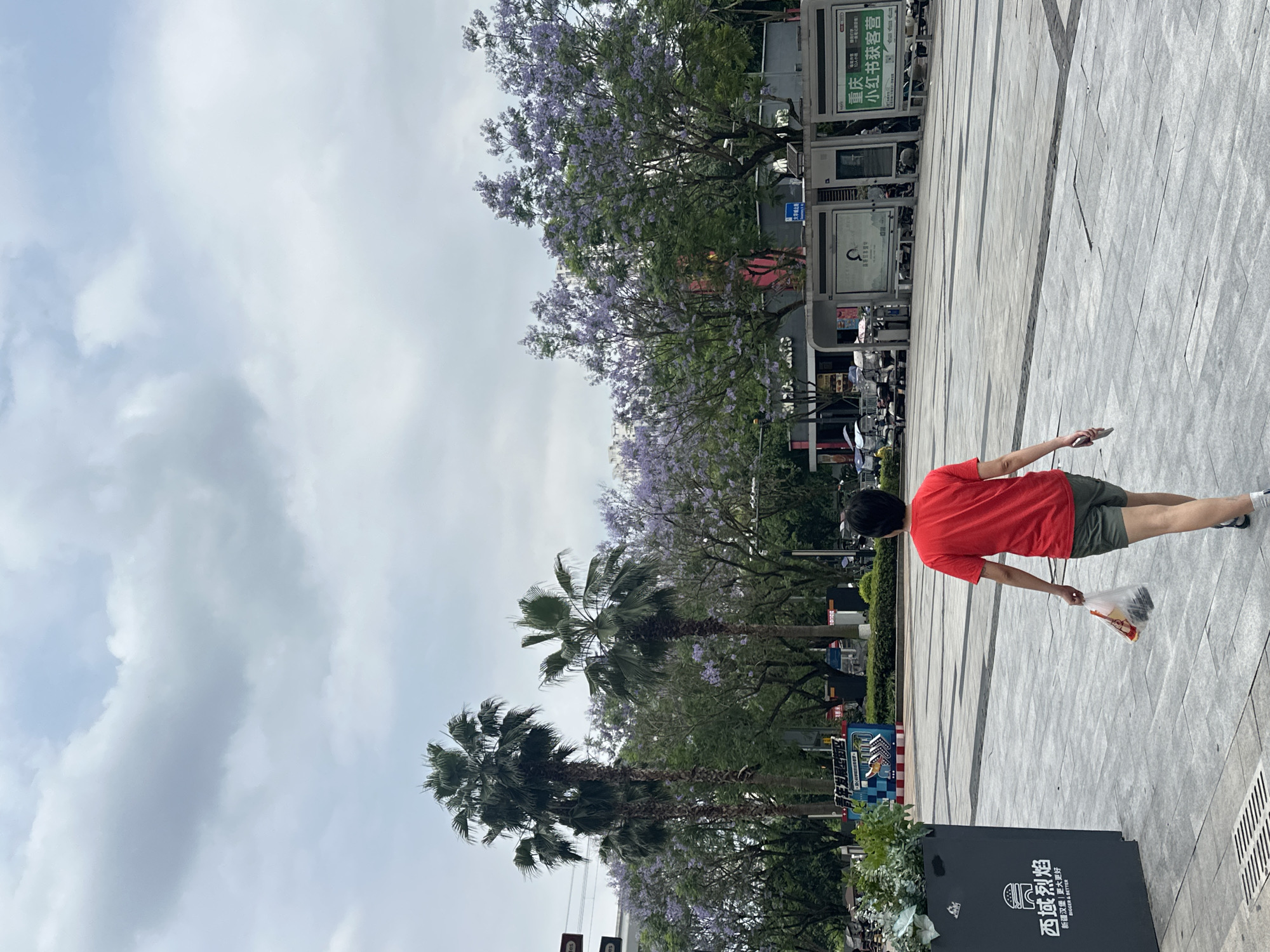 The image shows a man in a red t-shirt and green shorts walking on a paved sidewalk in Chongqing High-tech Zone, China. The sky is cloudy. Along the side of the sidewalk are palm trees and jacaranda trees in bloom, as well as advertisements and parking for bicycles. The sign shown is for a restaurant called "Western Flames", which serves Xinjiang burgers.