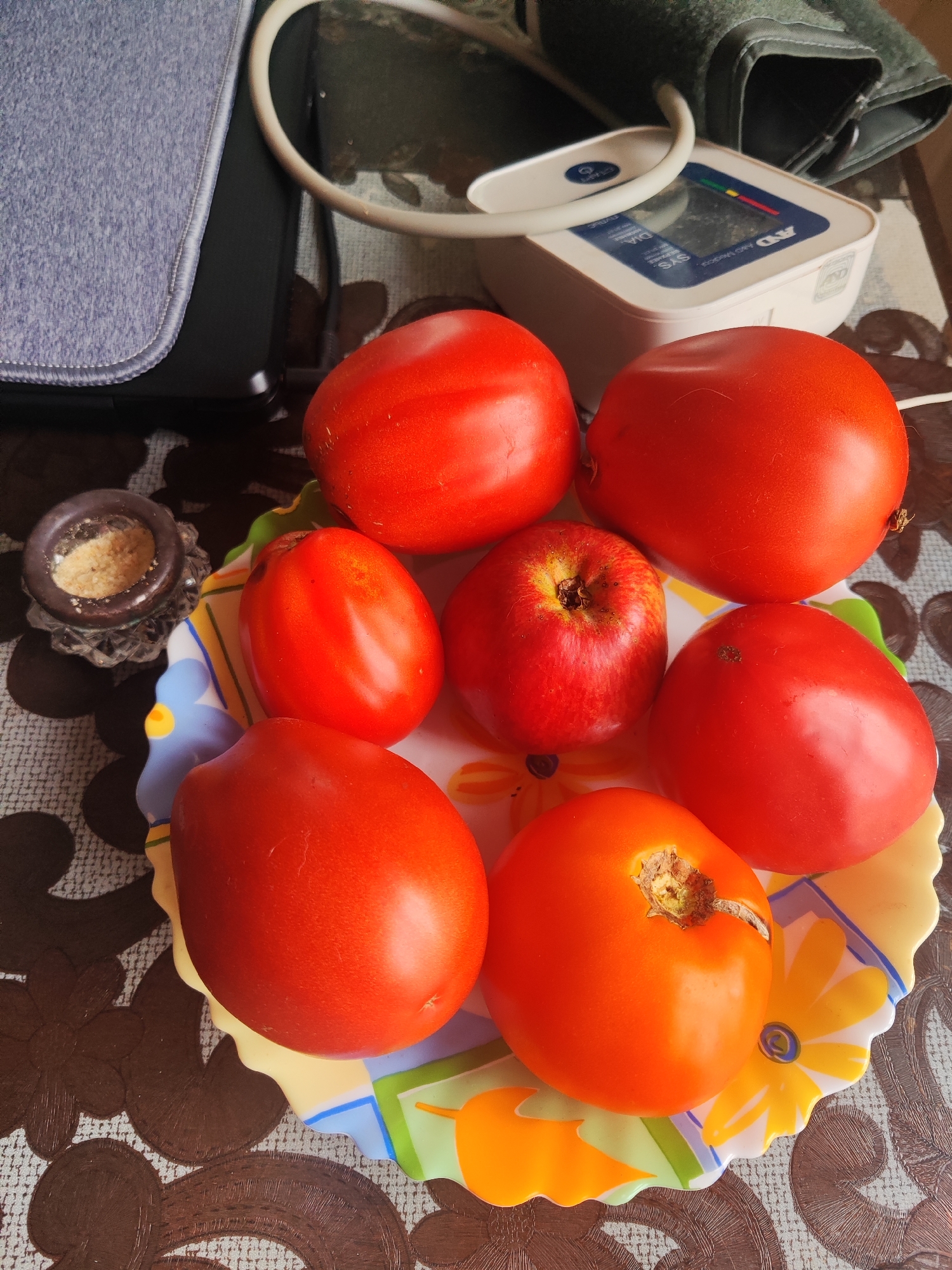 In Gvardeysk, Russia, on a patterned tablecloth, a colorful plate holds six red tomatoes and one red apple. A small, dark brown dish containing a light brown granular substance sits to the left of the plate. In the background, a white and gray blood pressure monitor with a cuff rests on the tablecloth. A dark gray object with a textured, purple-gray cover is positioned to the far left. The scene is illuminated by artificial light, casting shadows on the tablecloth.