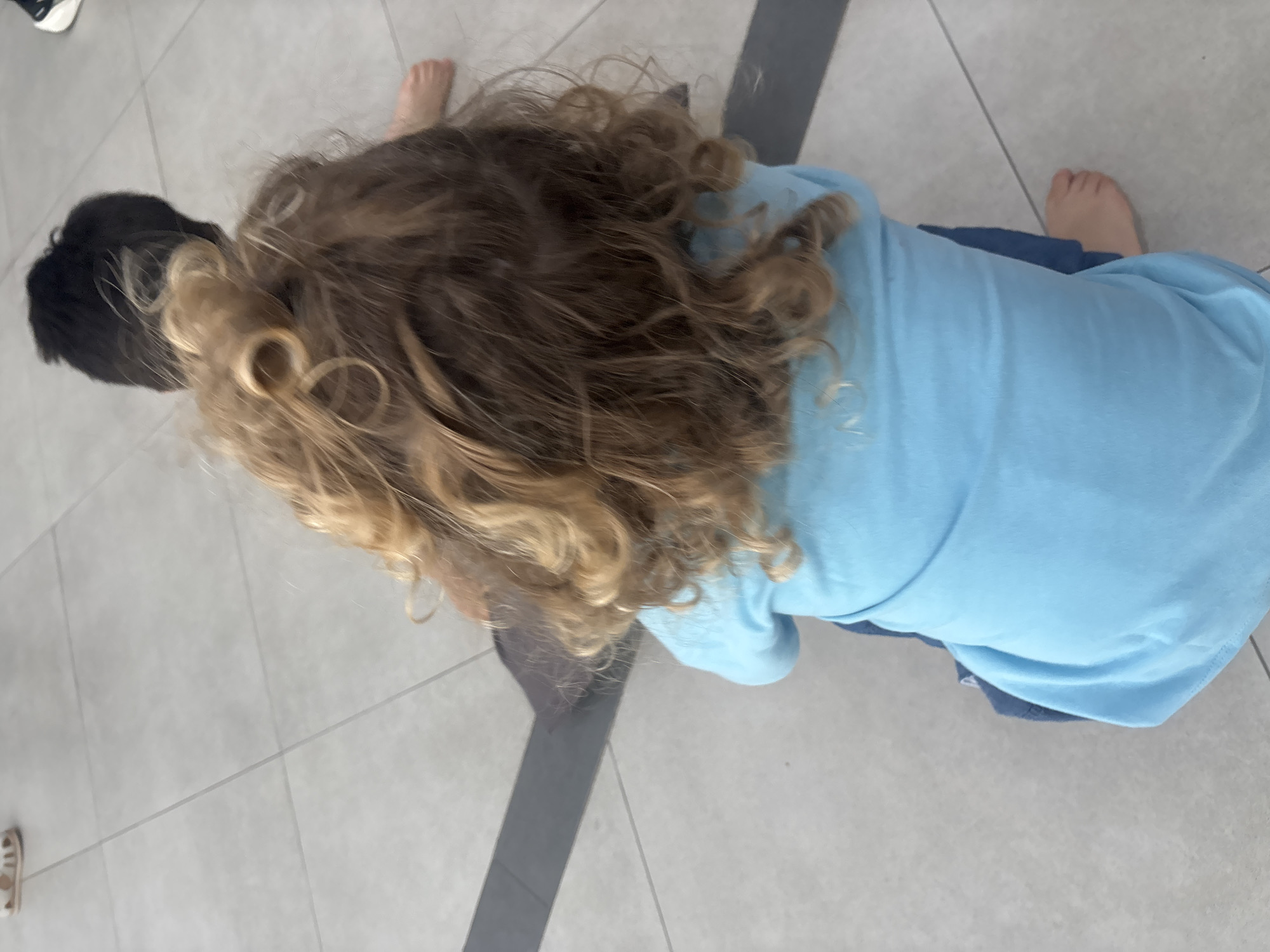 Two children are sitting on a tiled floor. The child in the foreground has curly blonde hair and is wearing a blue shirt and blue pants. The child behind them has black hair and is wearing a dark shirt. The floor is made of light-colored square tiles with dark grout lines. A dark gray stripe runs diagonally across the floor in the background. There is a partial view of a white sandal and a black shoe on the floor in the upper and right portions of the image, respectively. The scene appears to be indoors in a public space. There is no information in the image to indicate a specific location in Dubai, United Arab Emirates.