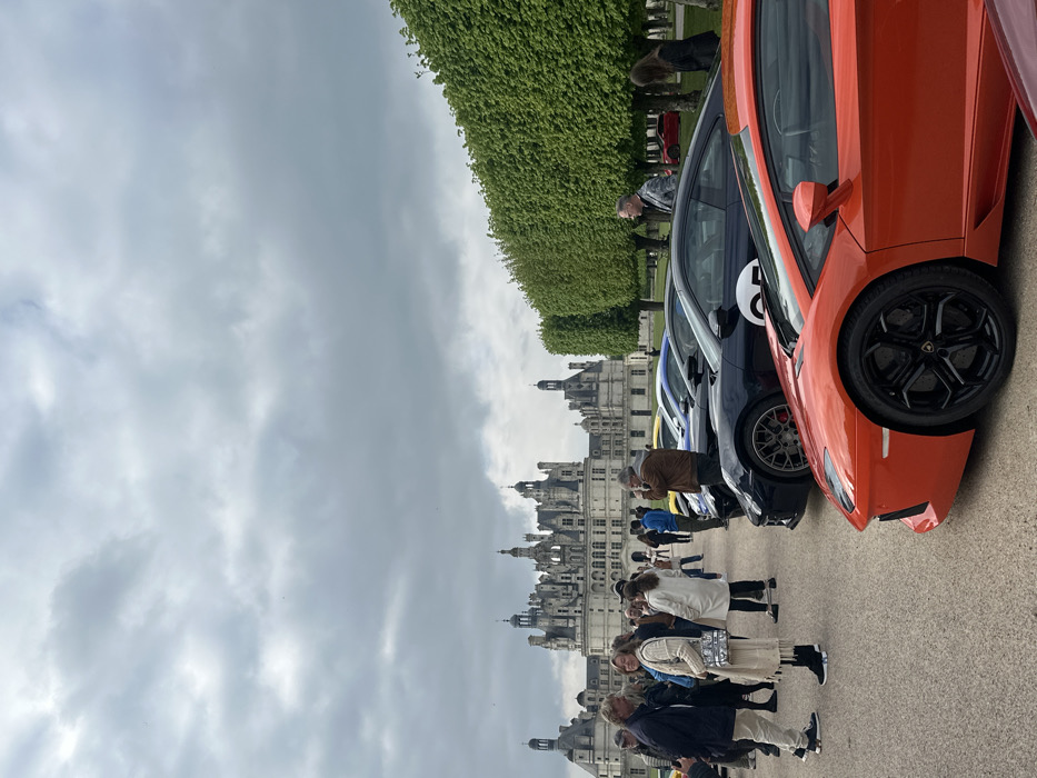 The image shows a group of people standing in front of a row of luxury sports cars parked outside the Chateau de Chambord, located in Chambord, France (latitude: 47.615, longitude: 1.519). The people are dressed casually and appear to be admiring the cars. The cars are all high-end models, including a Lamborghini Aventador, a Porsche 911, and a Ferrari. The chateau can be seen in the background, towering over the scene. The sky is overcast with clouds, and the trees are in full bloom, creating a lush and picturesque setting.