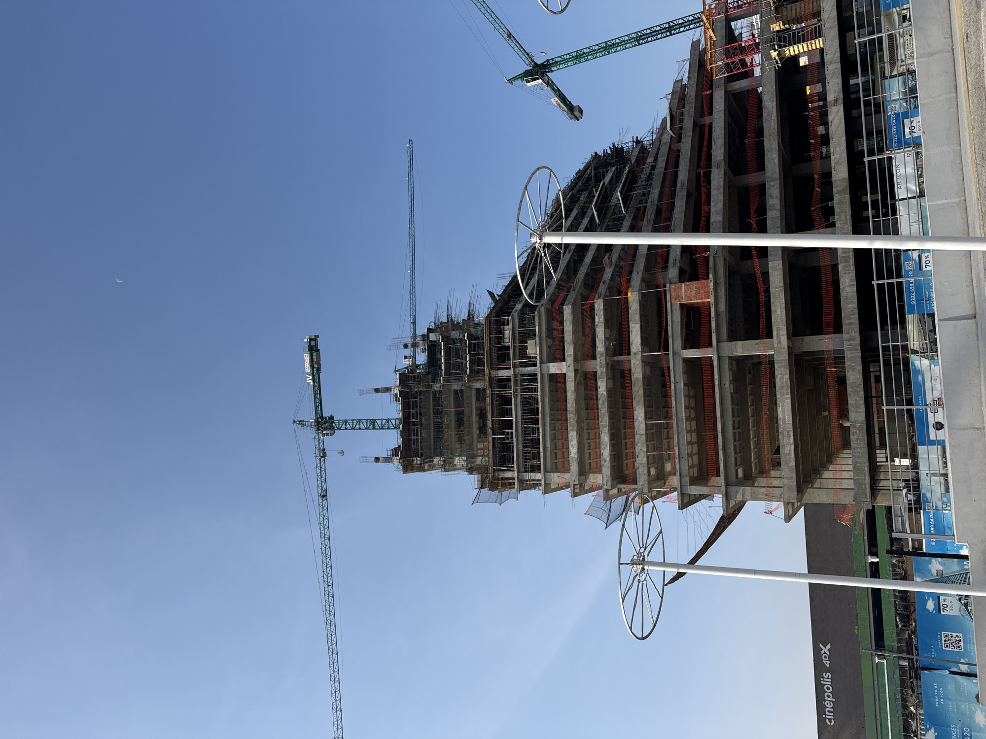 The image depicts a construction site for a high-rise building in Puebla, Mexico. The structure, which appears to be in the early stages of development, consists of a concrete framework with some orange safety netting. Several green construction cranes are visible in the background, positioned to aid in the building process. The sky is a clear blue, and the moon is visible. At the bottom of the image are light poles with large circular artistic decorations. A Cinépolis 4DX sign is present, suggesting the building is adjacent to or part of a commercial development that includes a cinema. The construction site appears to be near an advertising display indicating that 70% of the property is sold.