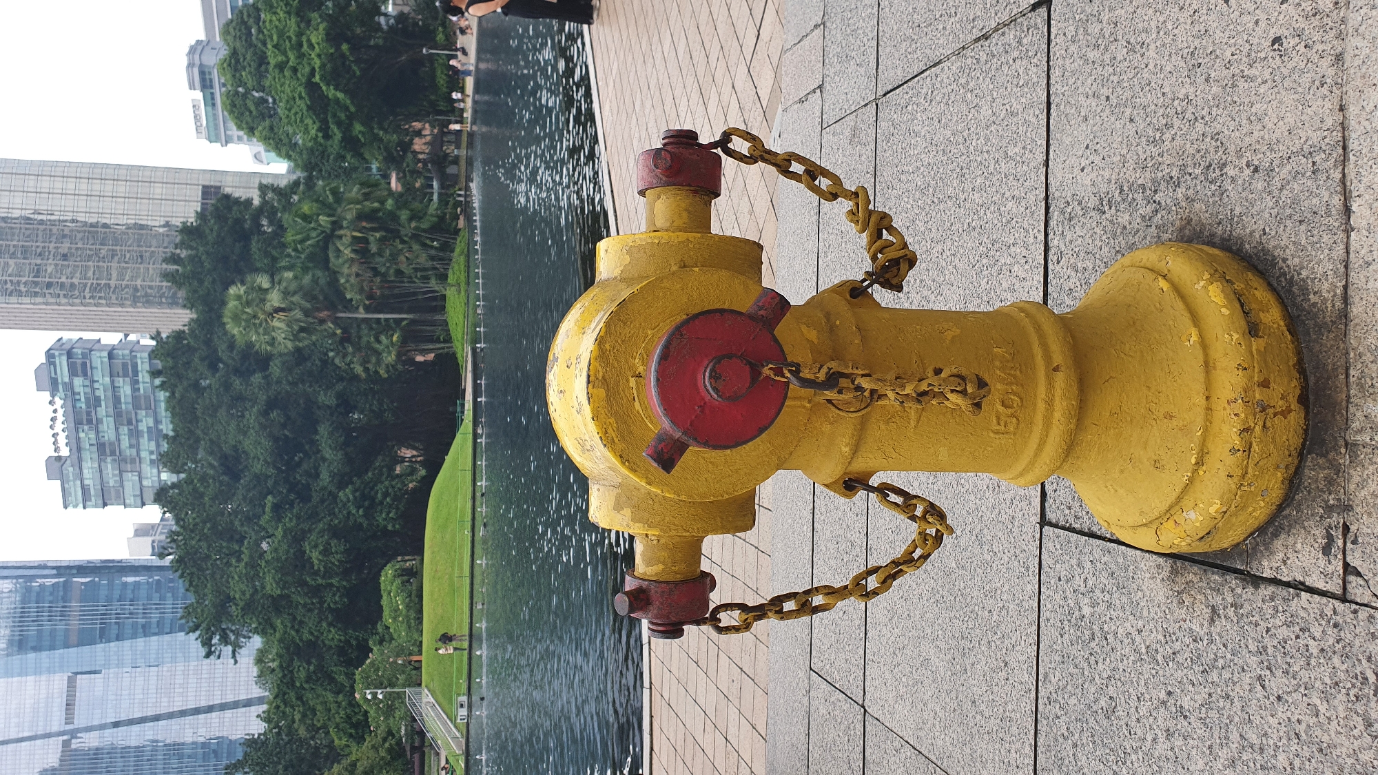 A bright yellow fire hydrant with red accents and rusty chains stands in the foreground on a paved walkway. Behind the hydrant is a large body of dark water, likely a public fountain or lake. In the middle ground, there's a green grassy mound with two individuals walking on it and a series of small fountains. The background features a dense cluster of trees, including palm trees, and several modern skyscrapers, suggesting an urban park setting in Kuala Lumpur, Malaysia. The overall atmosphere is one of a public space blending nature and modern architecture.