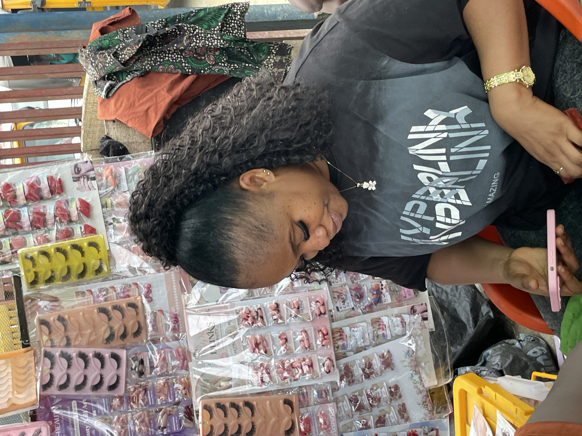 In the city of Lekki, Nigeria, a Black woman is seated, facing down and to the right, as she holds a pink mobile phone in her left hand. Her right hand is positioned over the phone, and her left hand holds a red object. The woman has long, curly black hair styled in a high ponytail and is wearing a black t-shirt with a large, white graphic design and a small, gold pendant in the shape of a teddy bear. She has on a gold wristwatch on her right wrist and a small gold stud earring in her left ear.
Behind the woman, a display of cosmetic products is visible. These include various sets of false eyelashes in pink and yellow packaging, and rows of small packages containing what appear to be artificial fingernails. Some of the packaging has floral designs. To the right of the cosmetic display, an orange fabric and a black, green, and white patterned cloth are draped. In the background, a yellow structure, possibly part of a vehicle, is partially visible behind a striped metal fence. A bright yellow plastic container is also visible in the lower left corner of the image.