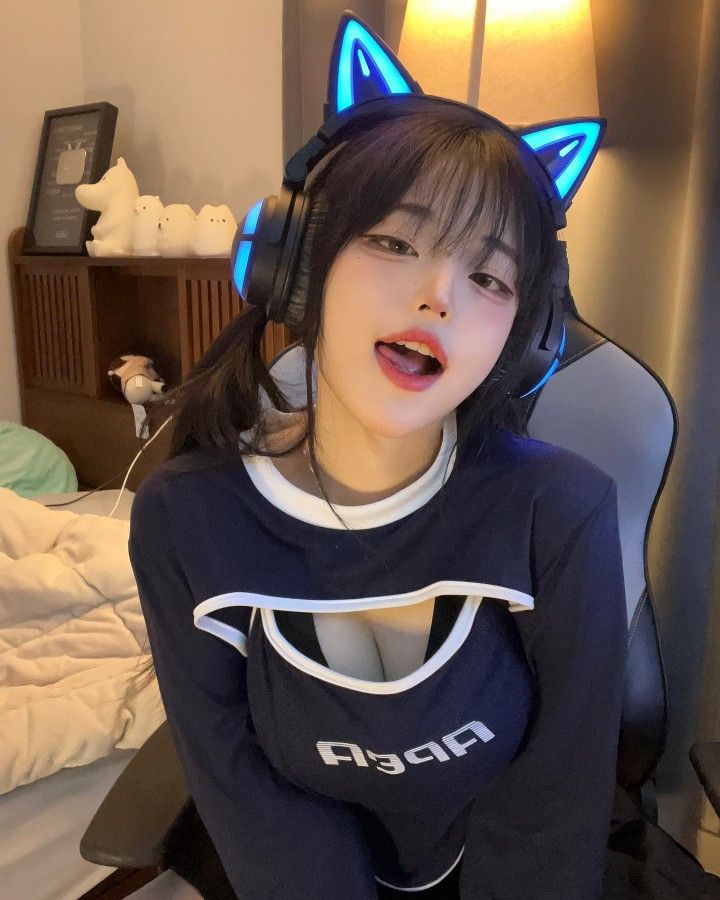 The image shows a young woman, likely in her late teens or early twenties, wearing a black and white long-sleeved shirt with a cutout and a pair of blue cat ear headphones. She is sitting in a chair, looking at the camera with her mouth slightly open. She is wearing a light-colored bra under her shirt, which is visible through the cutout. The chair she is sitting in appears to be a gaming chair with a black and gray color scheme. The background is a dimly lit room, with a bed to the left and a wall with some wooden paneling to the right. The image is taken at a close-up angle, making it difficult to discern any other details about the room. The woman's expressions and attire suggest a relaxed and informal atmosphere, possibly suggesting the setting is her home or a friend's house. The location of the image is in the city of Geneng, Indonesia, as indicated by the latitude and longitude coordinates (-7.45472, 111.427584).