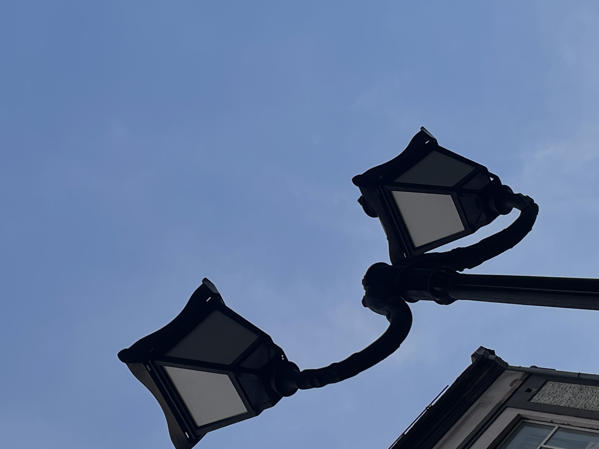 A low-angle shot captures a traditional double-headed street lamp against a clear blue sky. The lamp, rendered in silhouette, features ornate, dark metalwork supporting two lanterns with frosted glass panes. The structure of the lamp curves dramatically, with one arm extending upwards and to the left, and the other curving downwards and to the right. To the left edge of the frame, the top corner of an old building is visible, characterized by a grey facade and a white-framed window. This scene is located in the city of Rostov-on-Don, Russia.