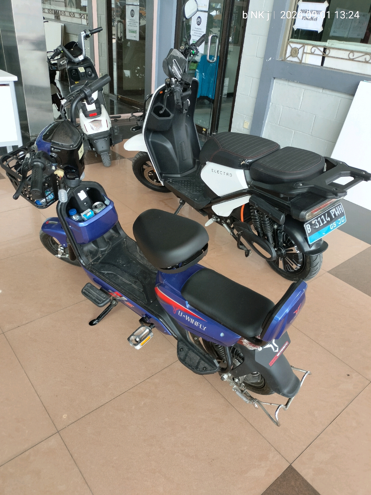 The image shows two electric scooters parked on a tiled floor in what appears to be an indoor area, possibly a lobby or showroom. The scooter in the foreground is blue with a black seat and a basket on the front. It has "U-WINFLY" written on its side. The scooter in the background is white with black accents and a black seat. It has "ELECTRO" written on its side and a license plate that reads "B 3114 PWH 05-26". The background shows a glass door with signs and a wall with grey and white tiles. A timestamp in the upper right corner of the image reads "2023 01 13:24". The presence of the license plate with Indonesian characters and the timestamp suggests the location is in Indonesia. Based on the provided location information, this scene is in Central Jakarta, Indonesia.