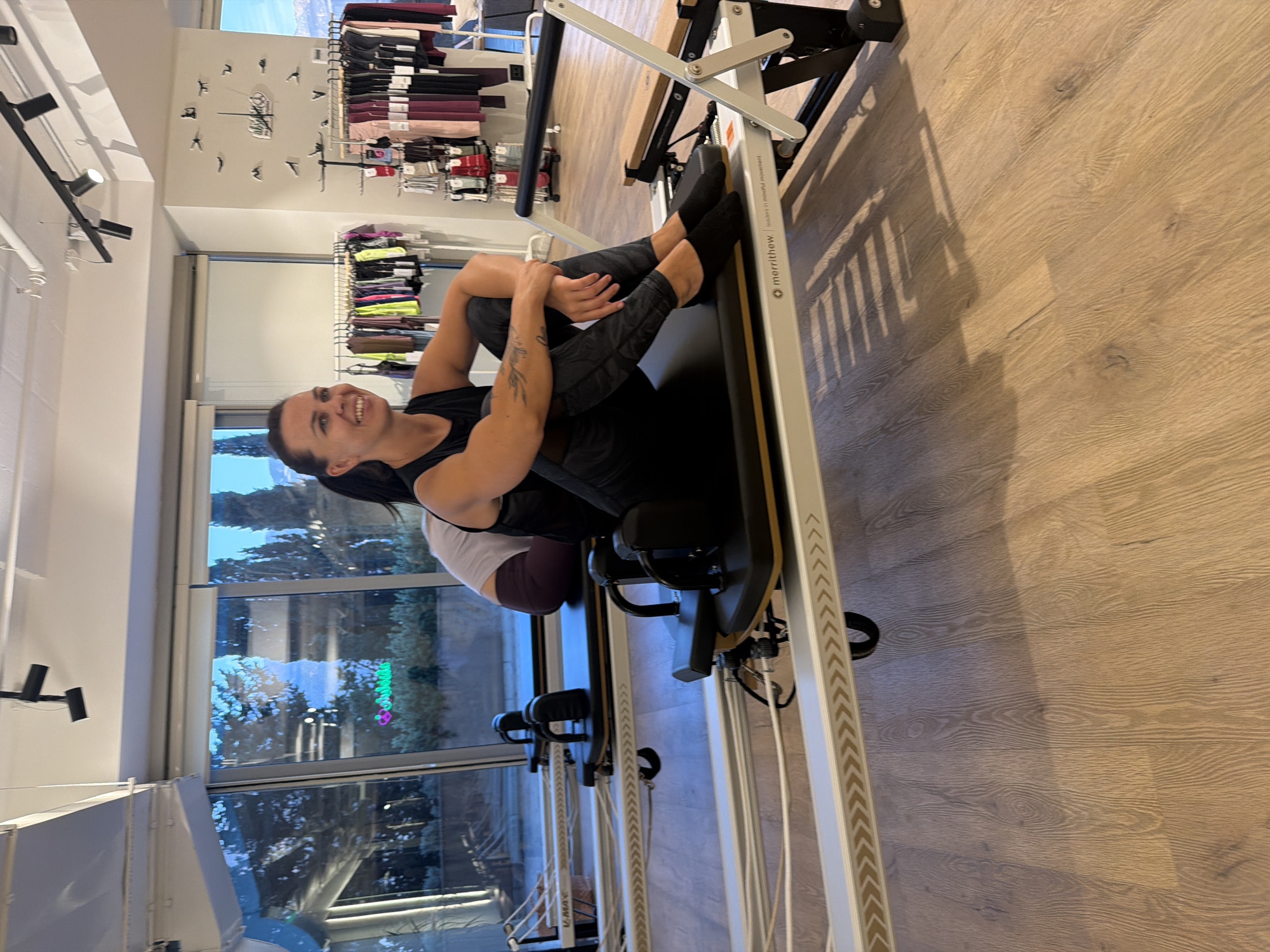 A woman is sitting on a piece of exercise equipment in what appears to be a fitness studio. The equipment is a reformer, common in Pilates studios, and has a black padded surface and metal rails. The woman is wearing a black tank top and patterned leggings, and black socks. She is smiling and looking towards the camera. In the background, there are more reformers, racks of athletic clothing, and large windows that look out onto an urban landscape with trees and mountains in the distance, suggesting the location is Boreti, Montenegro. The floor is made of light-colored wood.