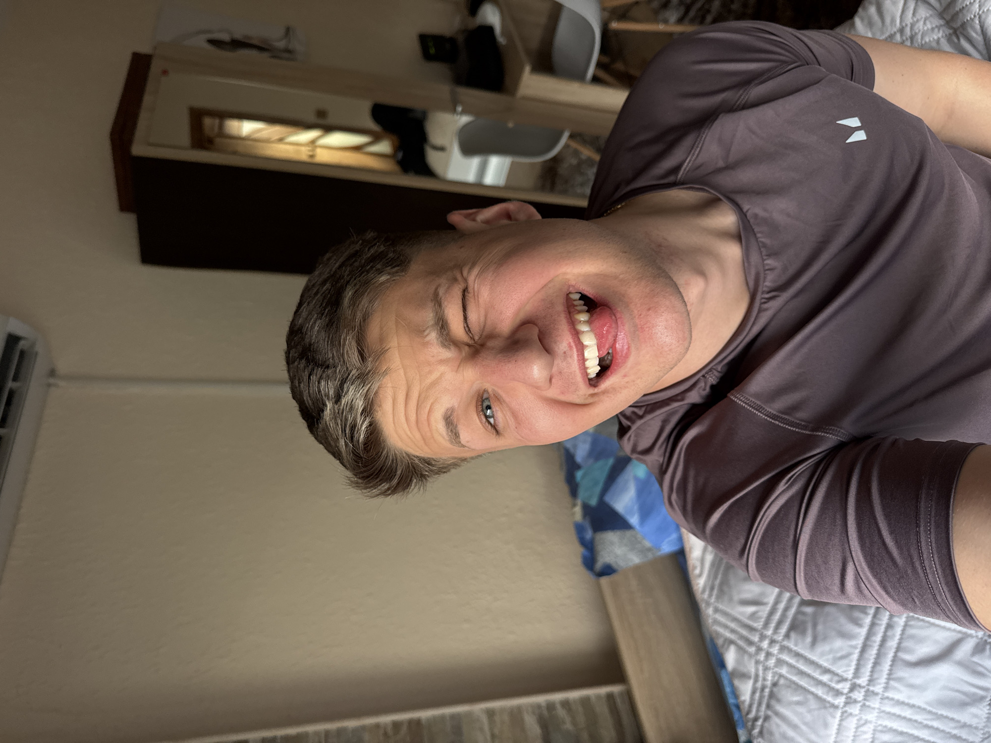 A man is taking a selfie indoors. He is making a funny face, winking one eye and sticking his tongue out. He is wearing a dark purple shirt and appears to be lying on a bed with gray and white bedding and a blue patterned blanket. In the background is a wall, an air conditioning unit, and a shelf with objects on it. The location is potentially a room in Burgas, Bulgaria, but this cannot be confirmed from the image alone.