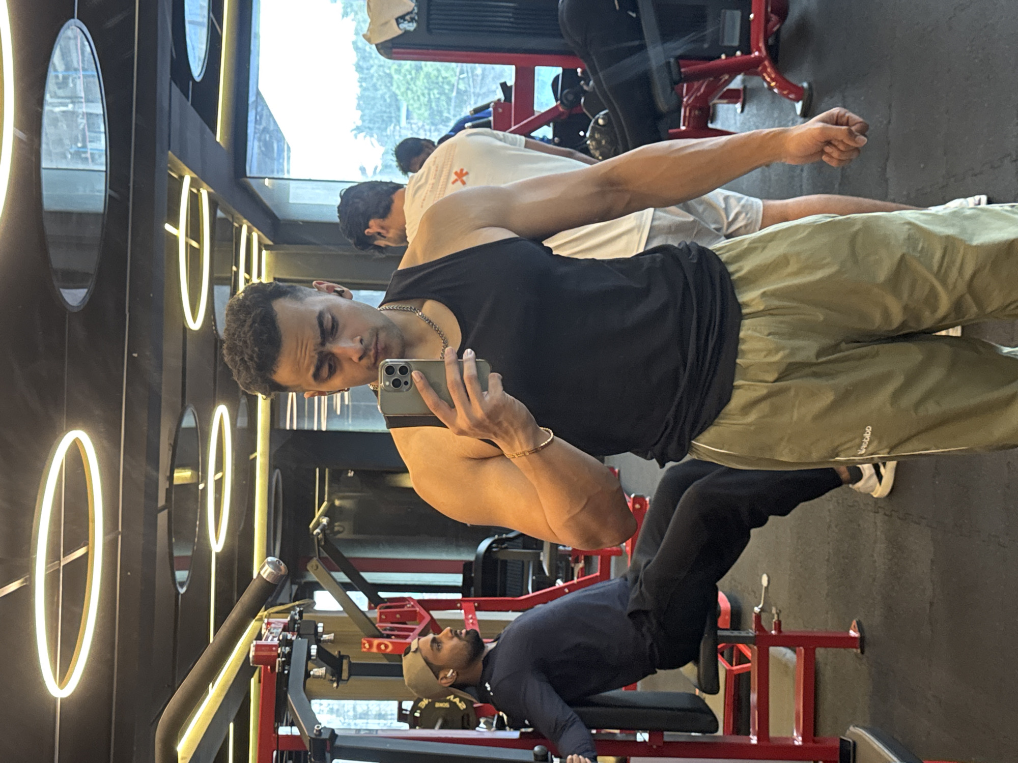 The image shows a man taking a mirror selfie in a gym. He is wearing a black tank top and light green athletic shorts. He has a chain necklace and a bracelet. In the background, another man is working out on a red gym machine, and two other individuals are visible further back. The gym appears modern with overhead circular lights and large windows. The setting suggests an urban environment.