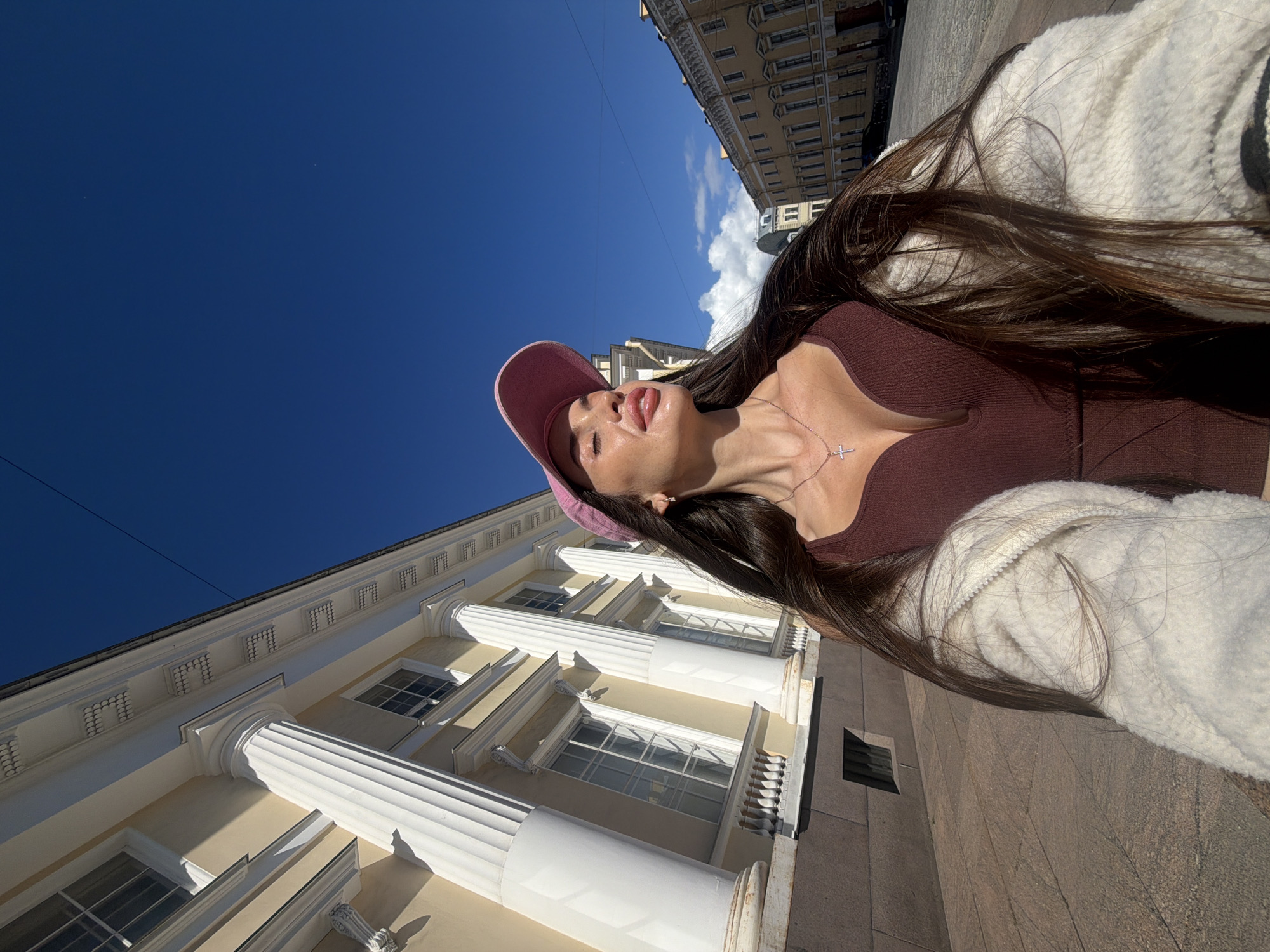 The image features a young woman with long, dark hair, wearing a pink baseball cap and a maroon top. She is also wearing a white fuzzy jacket. The woman is outdoors, with her head tilted back and eyes closed, appearing to bask in the sunlight. A delicate silver necklace with a cross pendant is visible around her neck.
The setting is an urban environment, characterized by classical architecture with large columns and multiple windows. The building's facade is painted a pale yellow, contrasted by white architectural elements. In the background, other buildings with similar architectural styles are visible, suggesting a historic city center. The sky above is a clear, bright blue with a few wispy clouds. The sun is high in the sky, casting distinct shadows, indicating it is daytime, likely midday.
The overall mood conveyed is one of relaxation and enjoyment of a sunny day. The composition, with the woman occupying a significant portion of the frame and the architectural elements serving as a backdrop, suggests a personal moment captured outdoors. No visible text is present in the image.