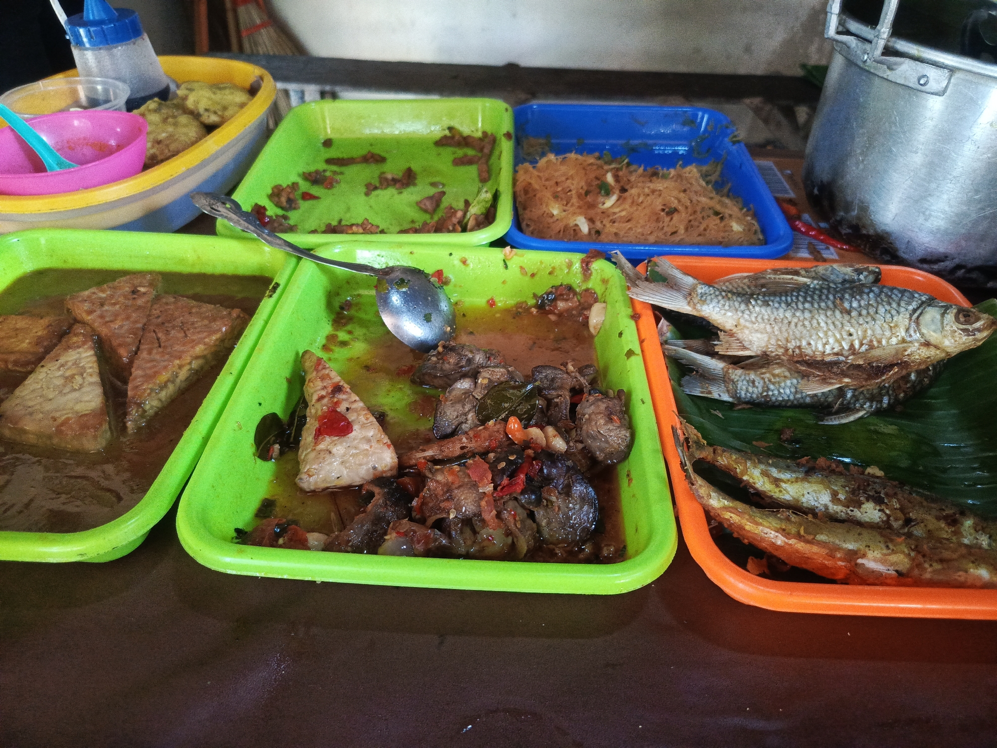 The image depicts a street food stall in Semarang, Indonesia, showcasing a variety of dishes. In the foreground, several green and orange plastic trays hold food items. One green tray contains what appears to be sliced tempeh in a savory liquid, while another green tray holds a spicy meat dish with chilies and other seasonings. An orange tray to the right displays two fried fish. In the background, a blue tray contains noodles, and a yellow oval container holds fried items, possibly tofu or tempeh. A pink bowl with a blue spoon is also visible. A large metal pot sits to the far right, suggesting cooking is taking place. The stall is set up on a brown surface, likely a table, with a glimpse of a wall and brooms in the background. The scene is indicative of a typical Indonesian food stall, offering a diverse selection of local cuisine.