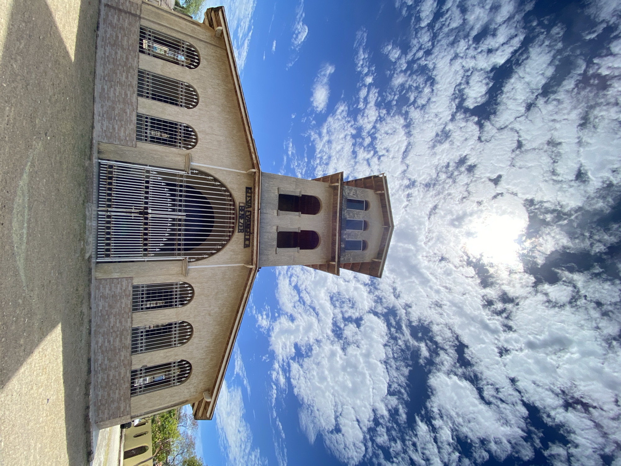 The image shows a building with a tower and windows. The building is beige and has a sign that reads "Iglesia Evangelica Eben-Ezer." There are metal bars covering the windows. The sky is blue with white clouds. The sun is shining brightly. This is likely the Iglesia Evangelica Eben-Ezer located in Juan Griego, Venezuela.