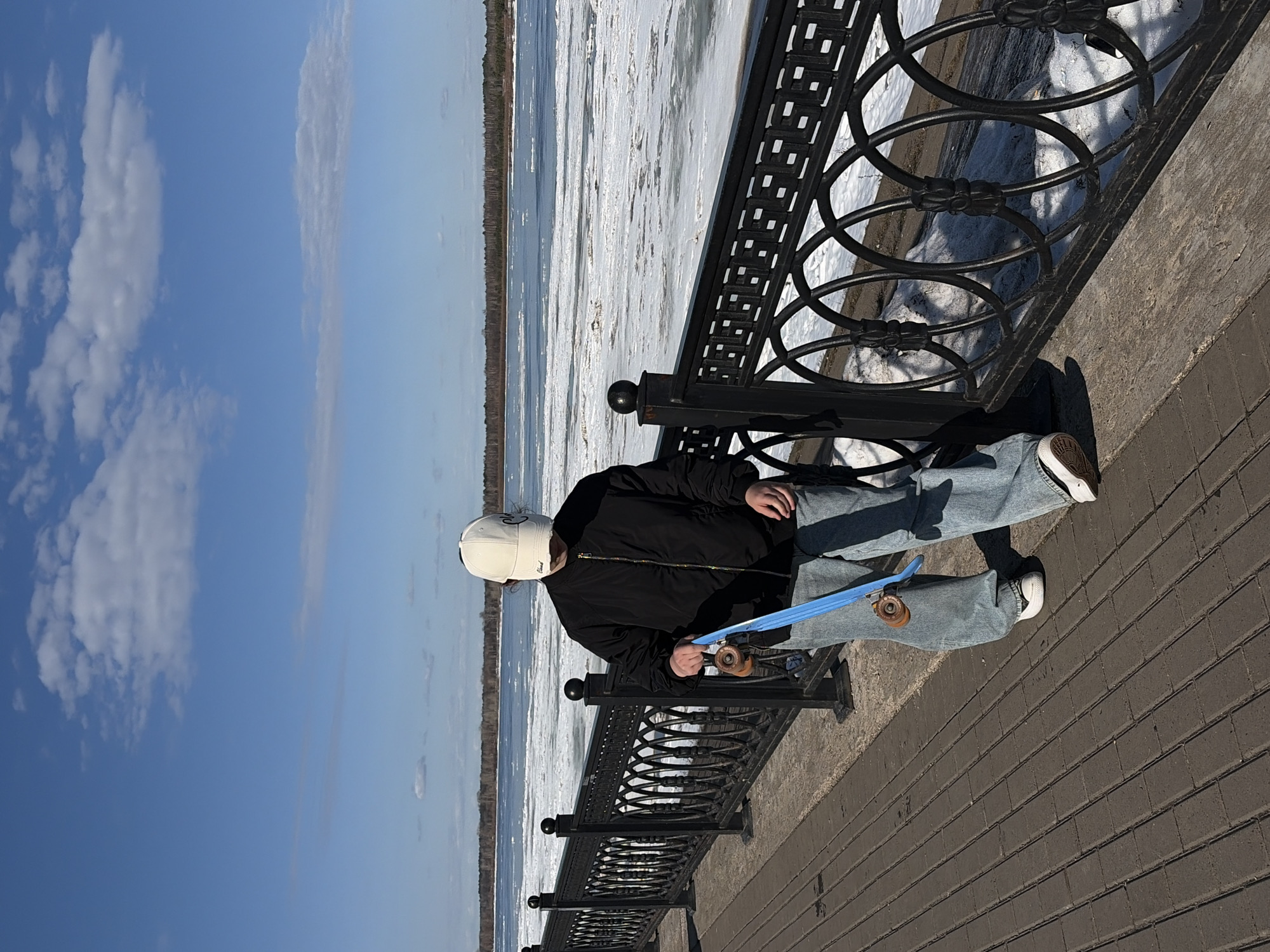 A person wearing a black jacket, light blue jeans, white shoes, and a white cap is standing on a brick walkway next to a black metal railing. They are holding a blue skateboard. Behind the railing, there's a body of water partially covered with ice, with a line of trees visible on the far shore. Above, the sky is blue with scattered white clouds. The location is Sarapul, Russia.