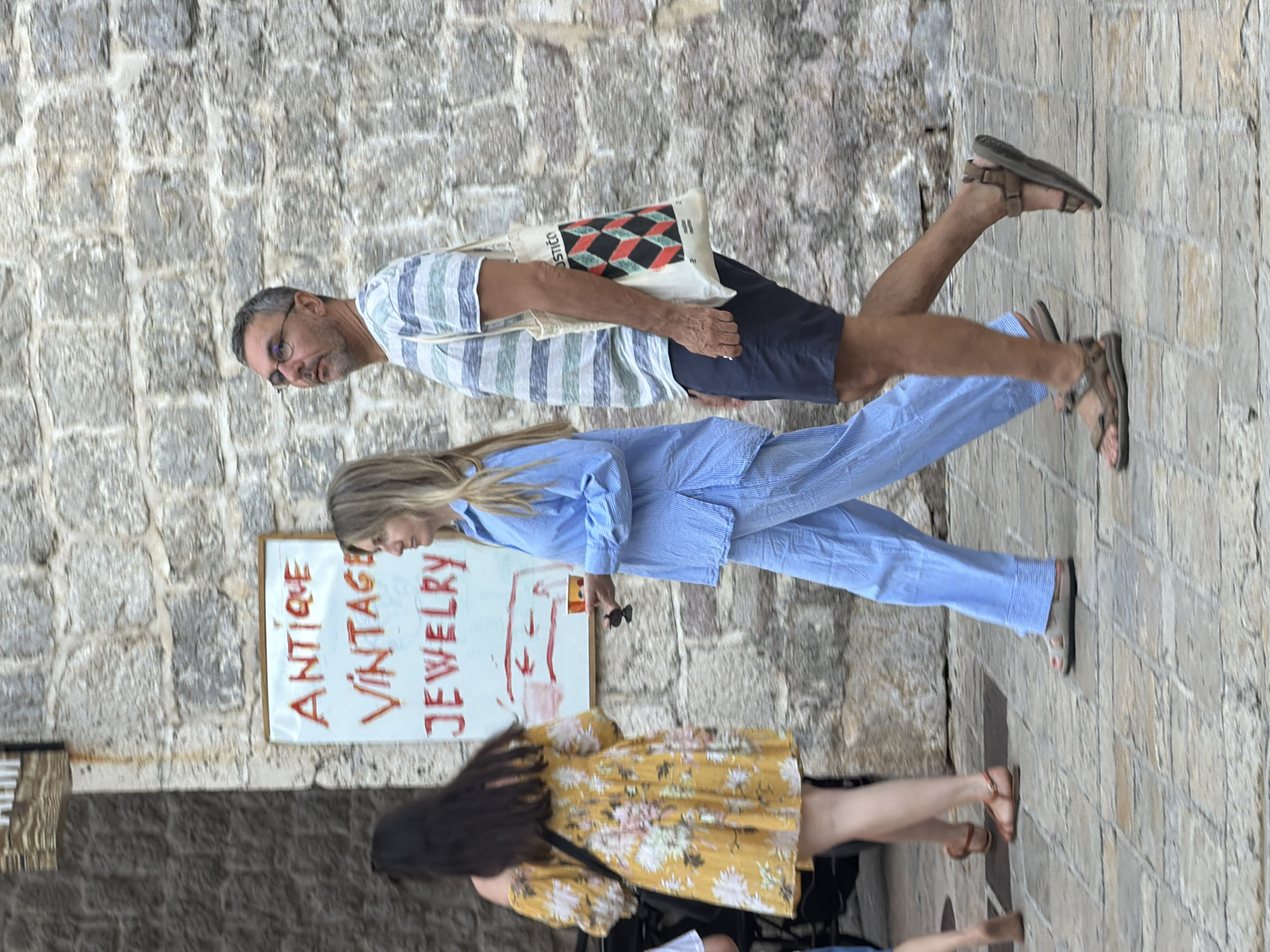 In Budva, Montenegro, on a cobblestone street with a stone building backdrop, a man and a woman walk past a sign that reads "ANTIQUE VINTAGE JEWELRY". The man, wearing glasses, a striped t-shirt, and dark shorts, carries a tote bag. The woman, in a light blue outfit, holds a small cup. Another woman in a yellow floral dress is partially visible to the left.