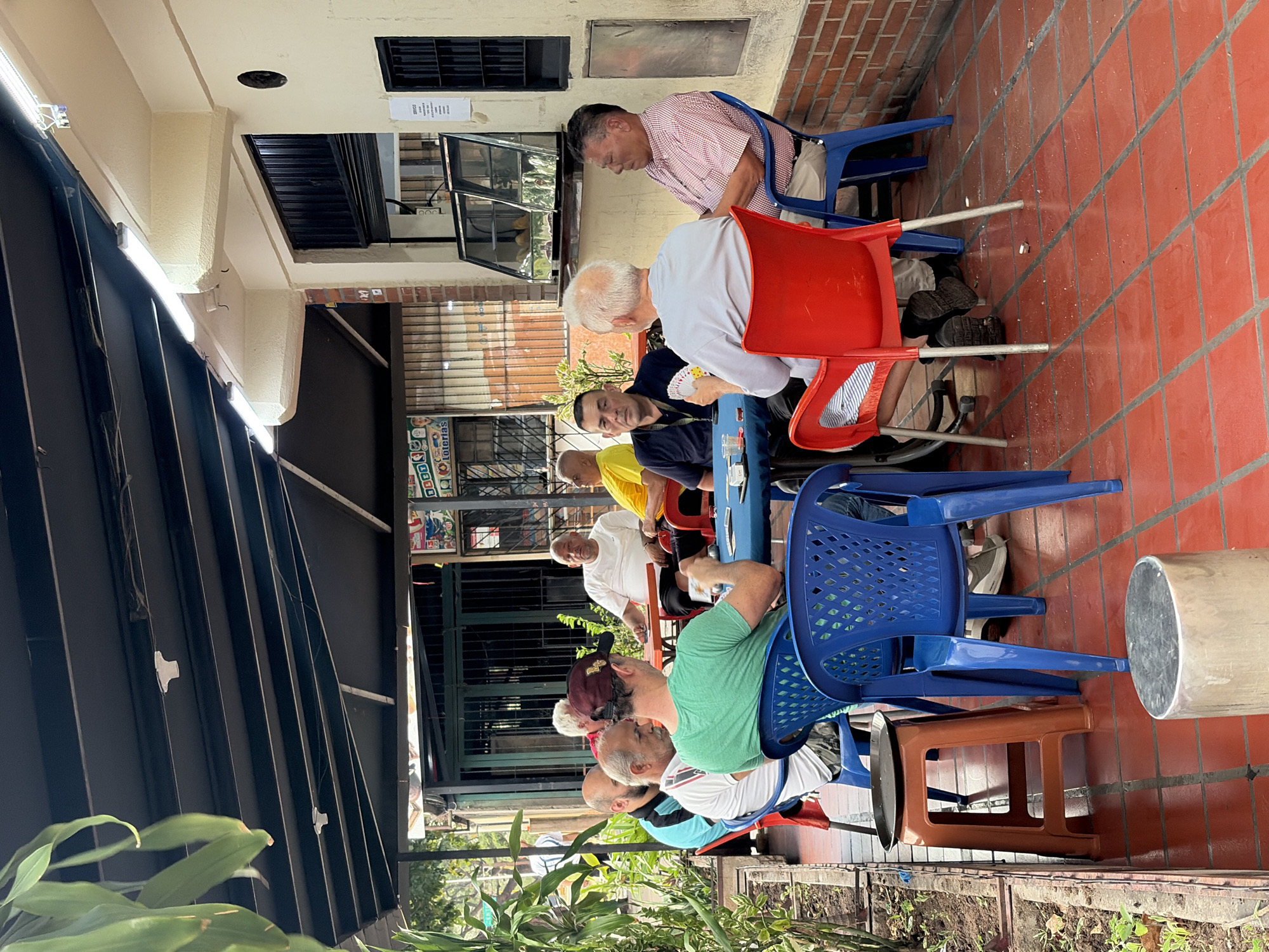 A group of men are sitting on chairs at a table in an outdoor setting. The scene appears to be under a covered walkway or patio area, with plants visible in the foreground. The men are dressed in casual clothing. Some of them are engaged in an activity, possibly a game of cards, as some have cards in their hands. The chairs are primarily blue and red plastic chairs. The location shows signs of being in a urban environment with buildings and shopfronts visible in the background. The red tile flooring is typical of many outdoor spaces in tropical climates. The image appears to be taken during daylight. Based on the clues, the location is in Ciudad Guayana, Venezuela.