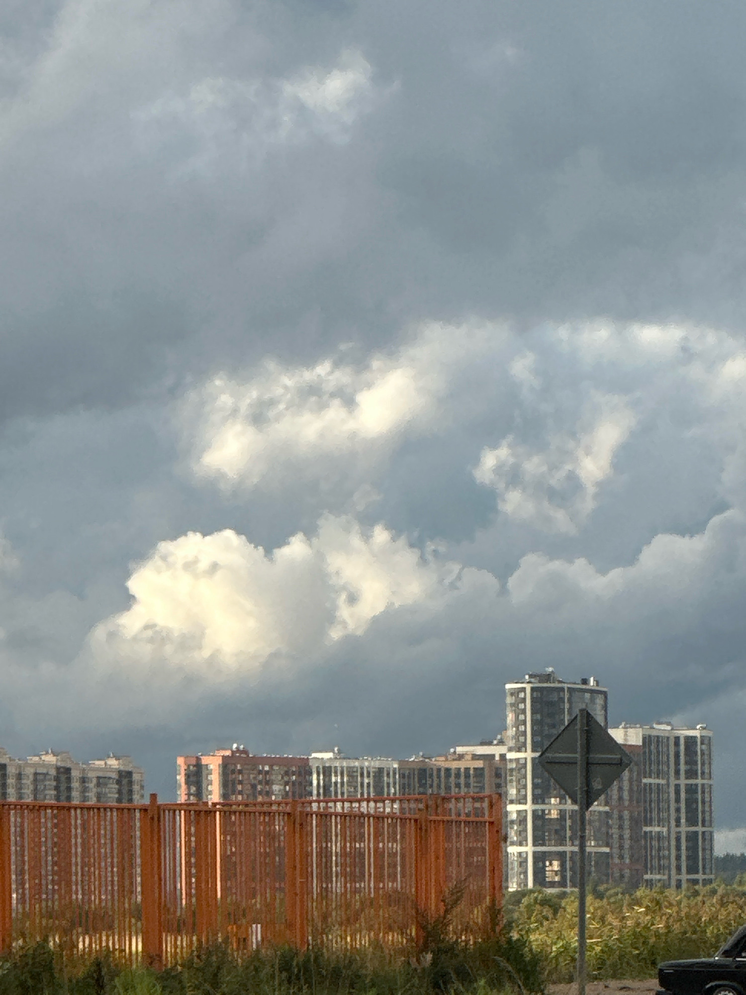 This is a photograph taken from a ground-level perspective, looking towards a cluster of modern residential buildings in Murino, Russia. The sky is overcast with dramatic, gray clouds, some of which are illuminated by sunlight breaking through, creating a contrast of light and shadow.
In the foreground, a bright orange metal fence runs horizontally across the frame, with sparse, dry vegetation visible behind it. Beyond the fence, several multi-story apartment buildings are visible. On the left, there are rows of light-colored buildings with numerous windows. In the center, a taller, modern building with a distinctive geometric design, featuring dark facades and white window frames, stands out. To its right, other buildings of varying heights and architectural styles are present, some with lighter facades and visible balconies.
A dark gray, diamond-shaped traffic sign on a pole is positioned in front of the central building. A small section of a black car is visible in the lower right corner of the image, partially obscured by vegetation. The overall impression is an urban landscape under a moody sky, with a focus on contemporary architecture.