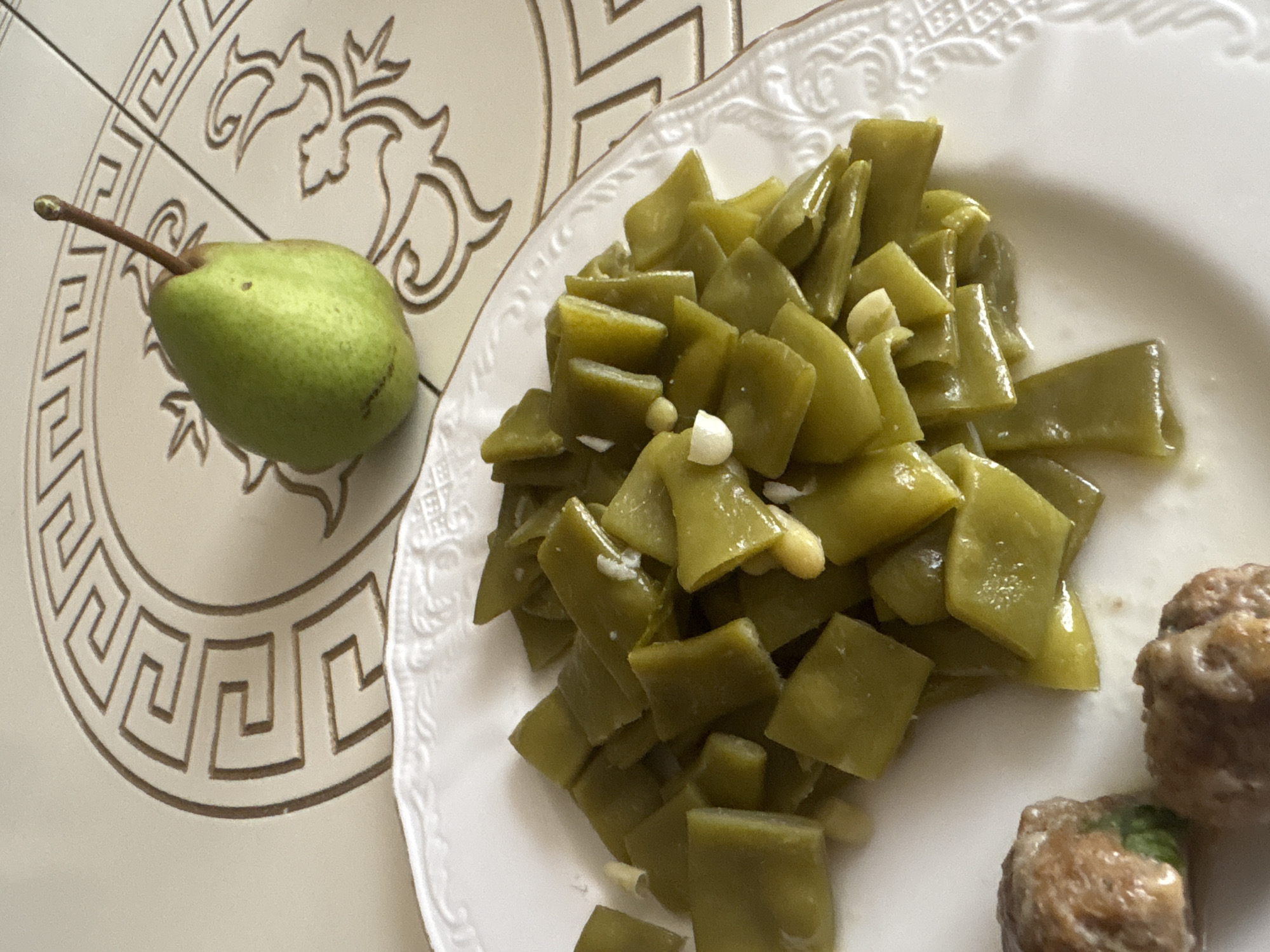 A close-up shot captures a meal served on a white patterned plate, with a green pear resting on a tiled surface in the background. The plate contains a generous portion of chopped green beans, some of which appear to be seasoned with garlic. Two meatballs are partially visible in the lower-left corner of the plate. The tiled surface features a decorative Greek key pattern. The image does not provide any information about the city of Georgiyevsk, Russia, or any people present in the scene.