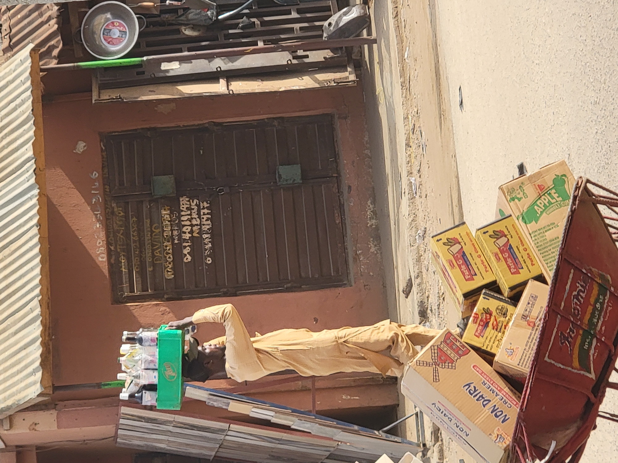 A man in a tan outfit stands near a shop with a closed, brown shutter, organizing bottles into a green crate. The shop's exterior is painted in shades of brown and orange, with handwritten text beside the shutter. To the man's right is a red cart filled with boxes, including those labeled "APPLE," "Parle-G," and "NON DAIRY CREAMER." The scene is likely in Maiburiji, Nigeria, based on the identified products.