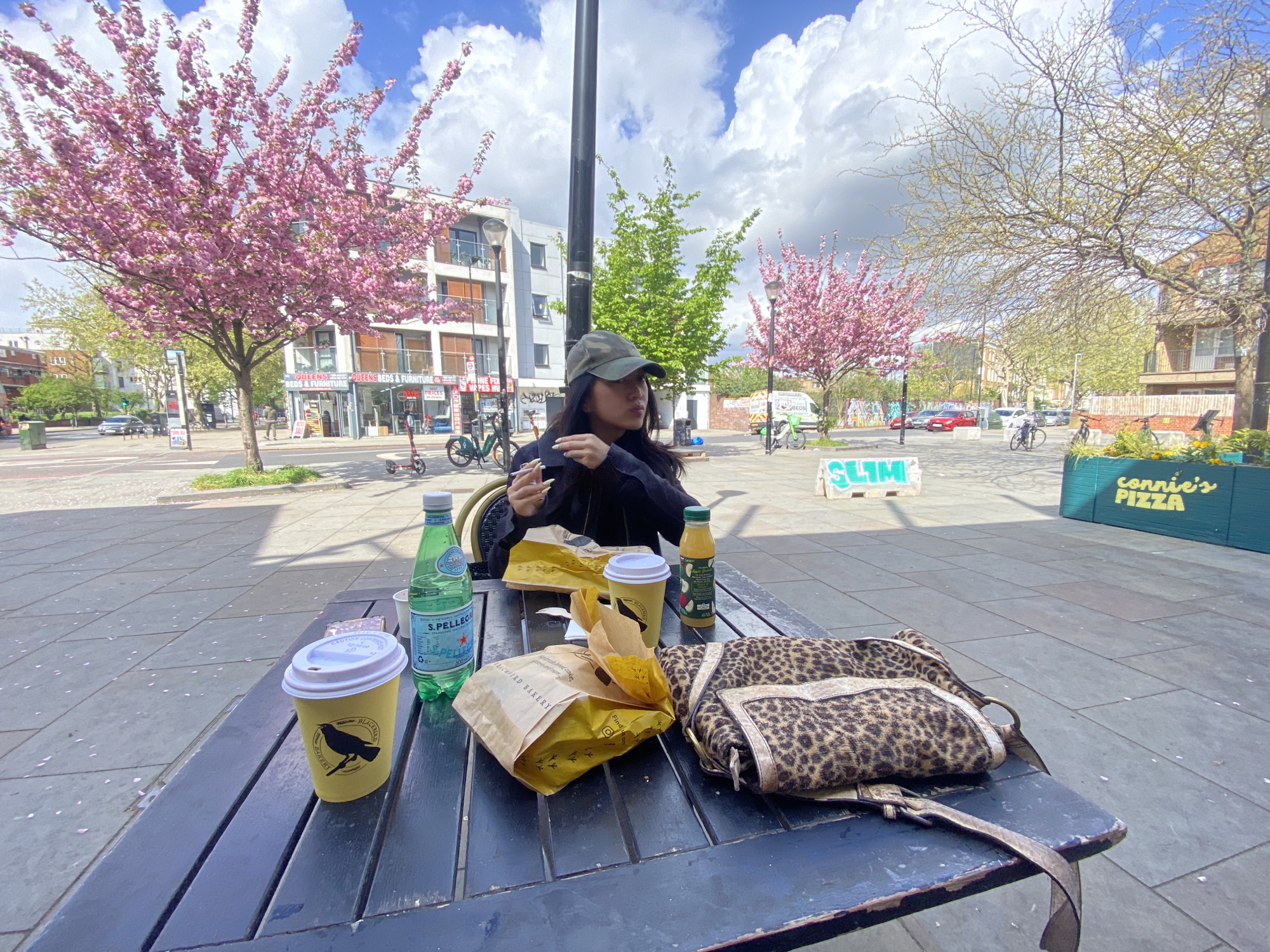 A person sits at an outdoor table, surrounded by various items. A yellow coffee cup with a bird logo, a bottle of S. Pellegrino, and two yellow paper bags labeled "BREADBIRD BAKERY" sit on the table alongside a bottle of orange juice and a leopard-print bag. The person is eating, and the atmosphere appears relaxed.
The backdrop includes a city street with buildings, trees with pink blossoms, and other street-level establishments, one of which is identified as "connie's PIZZA." A sign partially visible in the background reads "SLIM." The OCR identifies a store as "QUEENS BEDS & FURNITURE," further indicating the urban setting.