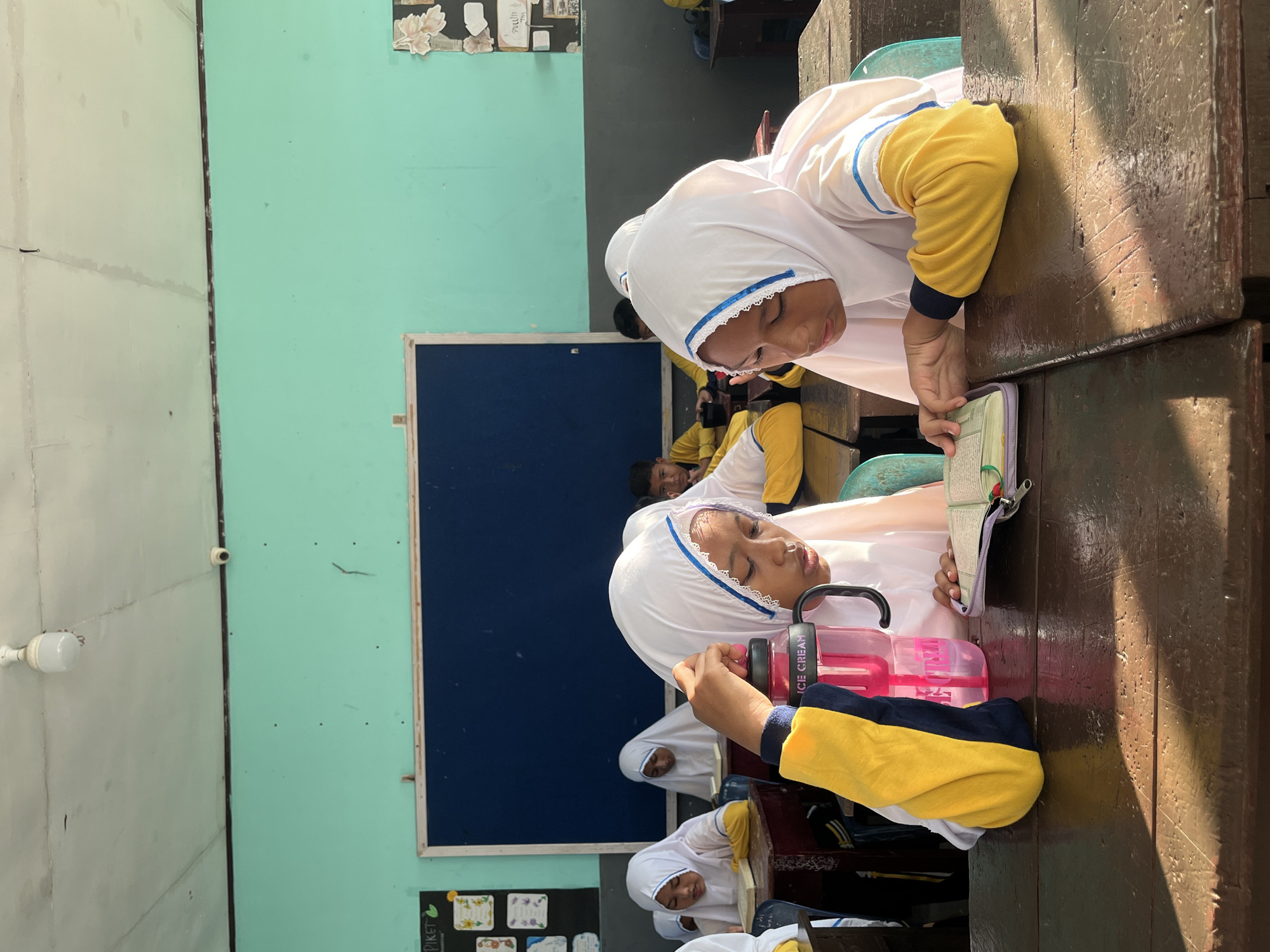 In a classroom in Lhokseumawe, Indonesia, two young girls wearing white hijabs and yellow and blue school uniforms are seated at wooden desks, engrossed in reading from an open book. The girl on the left holds a pink water bottle with "ICE CREAM" printed on it. The classroom has a light blue and green wall, a dark blue whiteboard, and a fluorescent light fixture on the ceiling. Sunlight streams in from the right, casting shadows across the desks. Other students are visible in the background, also appearing to be reading or studying.