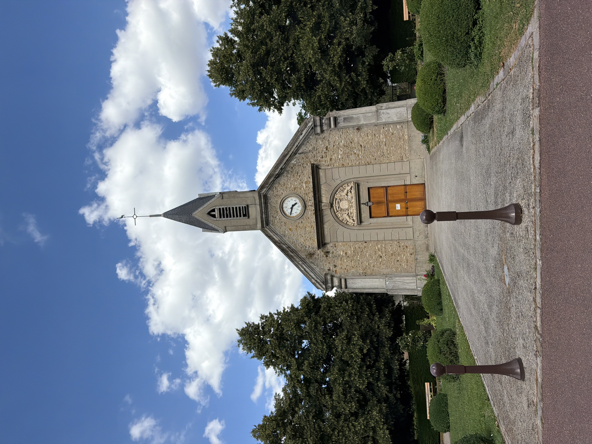 This is a low-angle, full-shot, exterior photograph of the Saint-Pierre church in Vélizy-Villacoublay, France. The church is constructed from stone with a dark steeple topped by a weather vane. A circular clock is visible on the facade above the wooden double doors. The church is set against a backdrop of a bright blue sky with scattered white clouds. Lush green trees flank the building, and manicured bushes and grass surround the entrance, which is accessed by a gravel pathway. Two dark brown bollards are positioned on the pathway in the foreground. Park benches are visible in the background on the left and right sides of the church. The overall impression is one of a serene, well-maintained religious building in a suburban or rural setting.