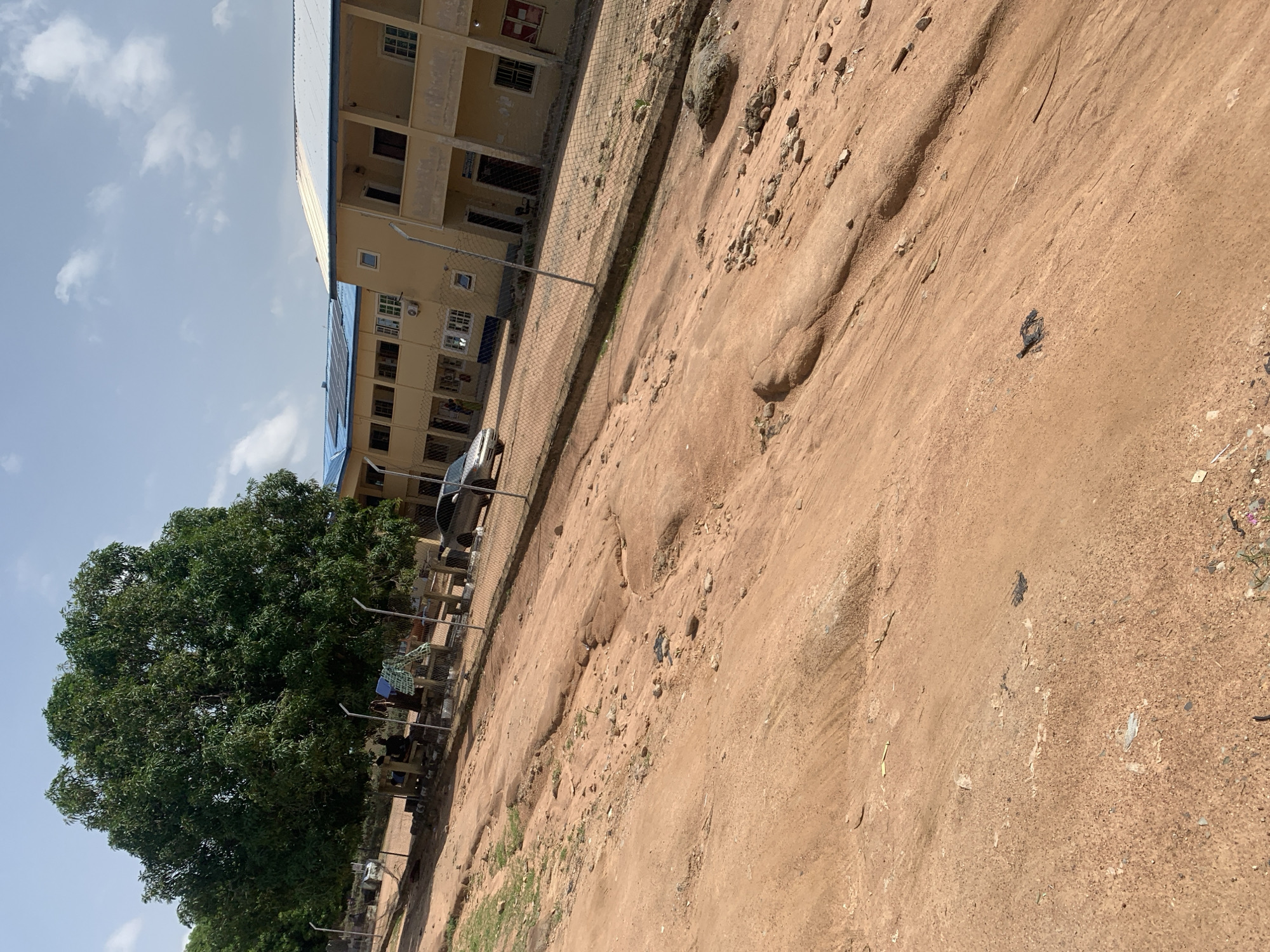 This image depicts a dusty, unpaved road in Jalingo, Nigeria. The road surface is uneven and shows signs of erosion and wear, with scattered small rocks and debris. To the left, a large, leafy green tree provides shade over a group of people seated on benches. A chain-link fence runs along the right side of the road, separating it from a two-story building complex. The buildings have yellow exteriors, tiled roofs (some blue, some white, with visible solar panels on one roof), and numerous windows and doors. A silver car is parked near the fence in front of the buildings. The sky is partly cloudy. The scene appears to be an outdoor area of a school or institutional facility within the city.