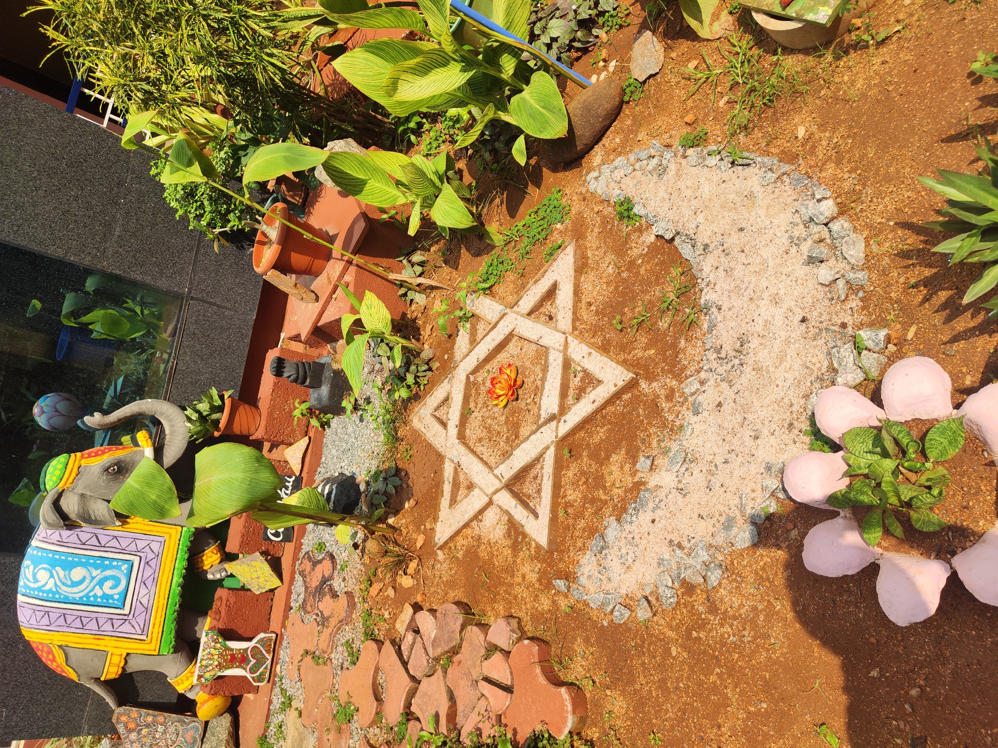 The image depicts a garden scene, possibly a private residence, in an unknown city and region. The ground is covered in reddish-brown soil.
Centered in the image is a star-shaped design. It is constructed of pale stone or concrete sections. A flower sits in the star's center. Adjacent to the star is a crescent shape formed from light-colored sand or gravel, bordered by small gray stones.
Several potted plants and various types of greenery surround the star and crescent designs. A painted elephant statue with a colorful saddle stands on the left side of the image. Reddish-brown pavers are also visible. A portion of a building is on the left side of the picture.