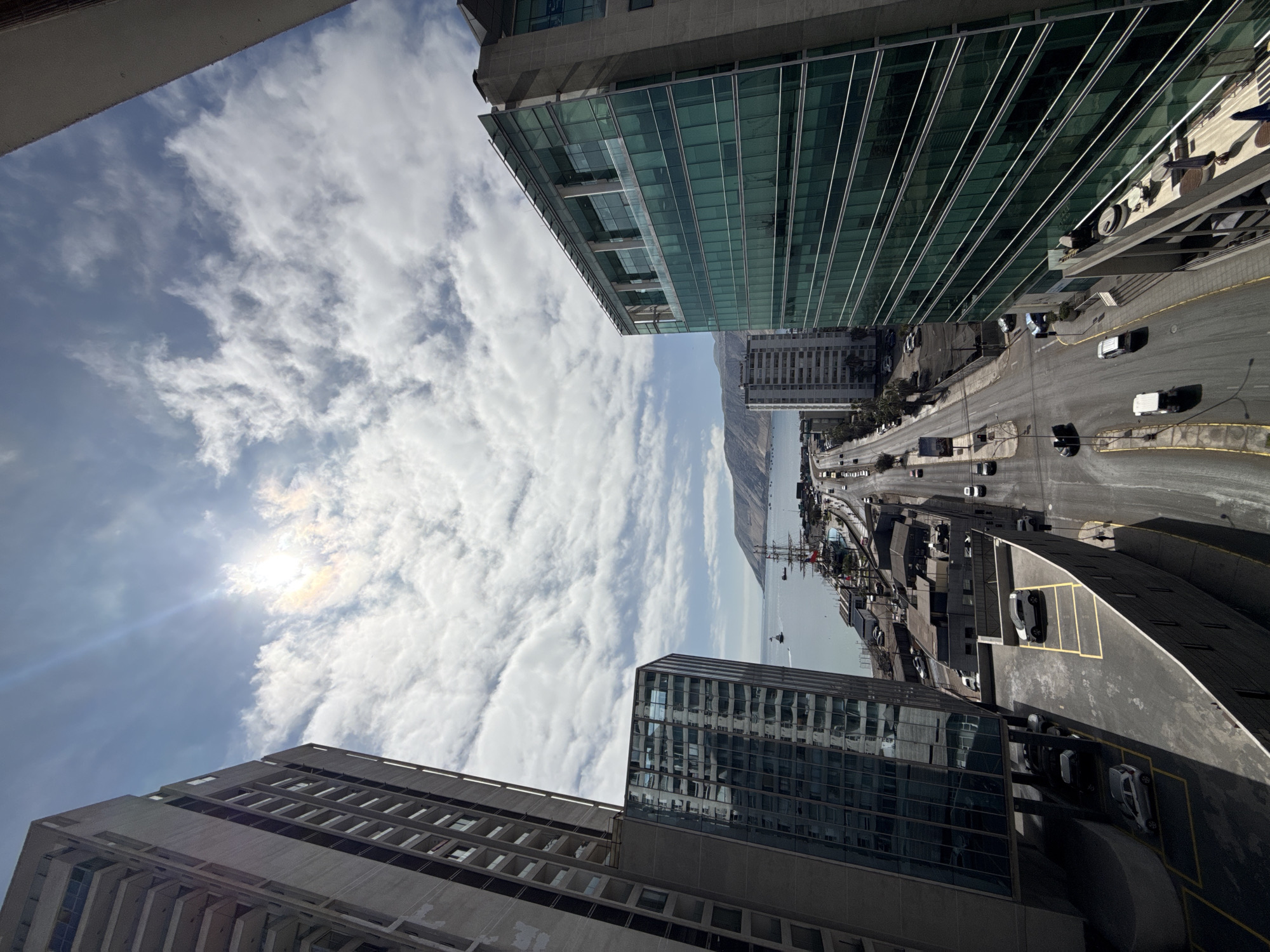 The image provides an elevated, wide-angle view of a coastal city with modern architecture and a harbor. The foreground is dominated by two tall, contemporary buildings; one on the left is a concrete structure with visible balconies and window frames, while the building on the right features a sleek, green-tinted glass facade. A multi-lane road runs parallel to the harbor, with several vehicles in motion and parked along its sides. The harbor itself is active, with various vessels, including what appears to be a large sailing ship docked near the shore. Beyond the harbor, a body of water stretches to the horizon, meeting a mountainous landscape under a bright, partly cloudy sky with the sun prominently visible. The scene suggests an urban environment in Iquique, Chile, given the characteristic coastal mountain range and the presence of a significant port.