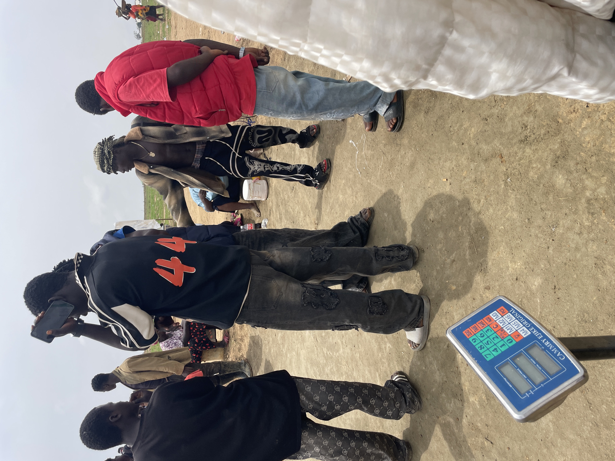The image shows a group of people standing on a flat, sandy surface, possibly in Maiyanga, Nigeria. Visible are a digital scale, legs and torsos of individuals in various clothing styles, including a red puffer jacket, black shirts, patterned pants, and ripped jeans. One person is holding a phone. The background suggests an open, outdoor environment.