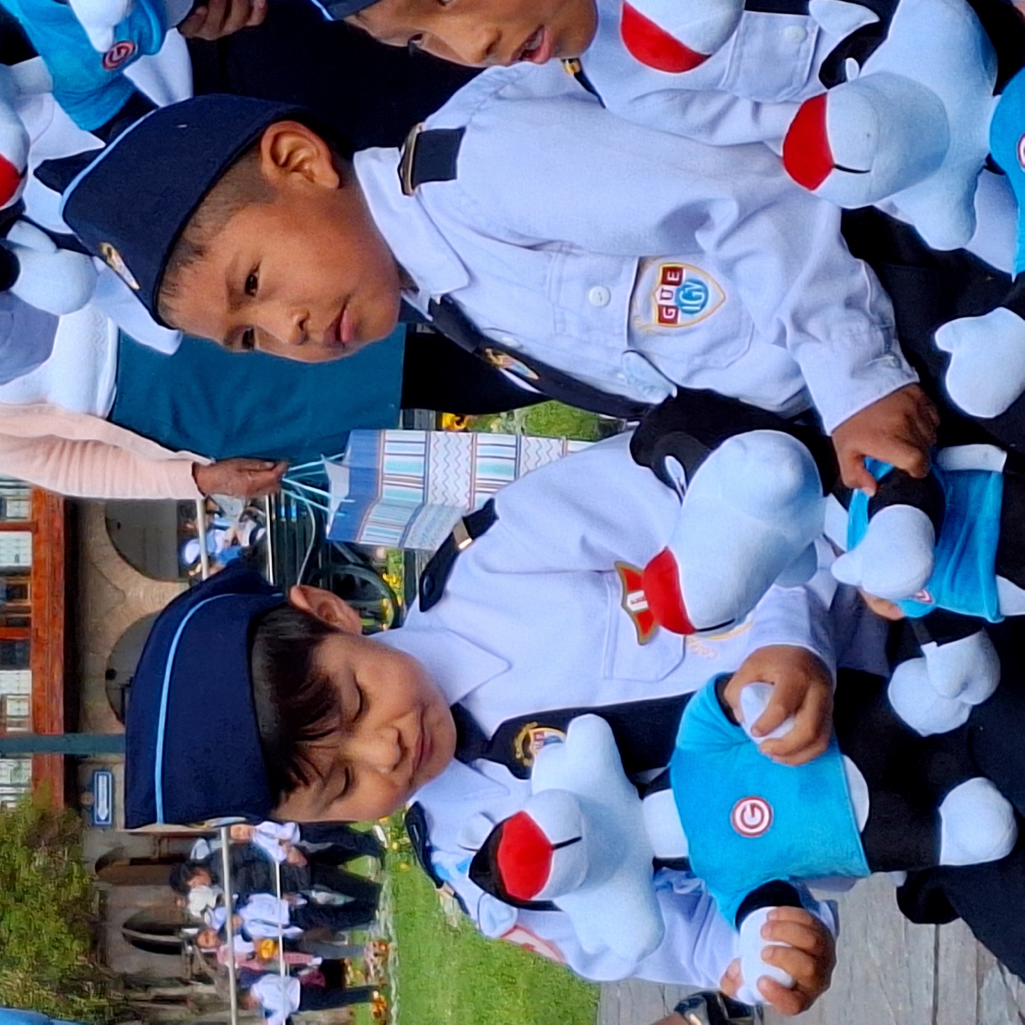 The image depicts a group of children in uniform, possibly students, gathered outdoors in what appears to be a city square or park in Cuzco, Peru. The children are wearing matching white shirts, dark ties, and dark caps with blue trim. Many of them are holding or interacting with stuffed toys. In the foreground, two boys are prominently featured. The boy on the left is looking down at a blue stuffed toy with a red and white circular logo. The boy on the right is looking off to the side, and his hands are on a similar stuffed toy. In the background, other children and adults are visible, along with buildings with arched doorways and a tall lamppost. A colorful gift bag with blue and white stripes is also present in the background. The overall scene suggests a public event or gathering, possibly related to a school activity or celebration.