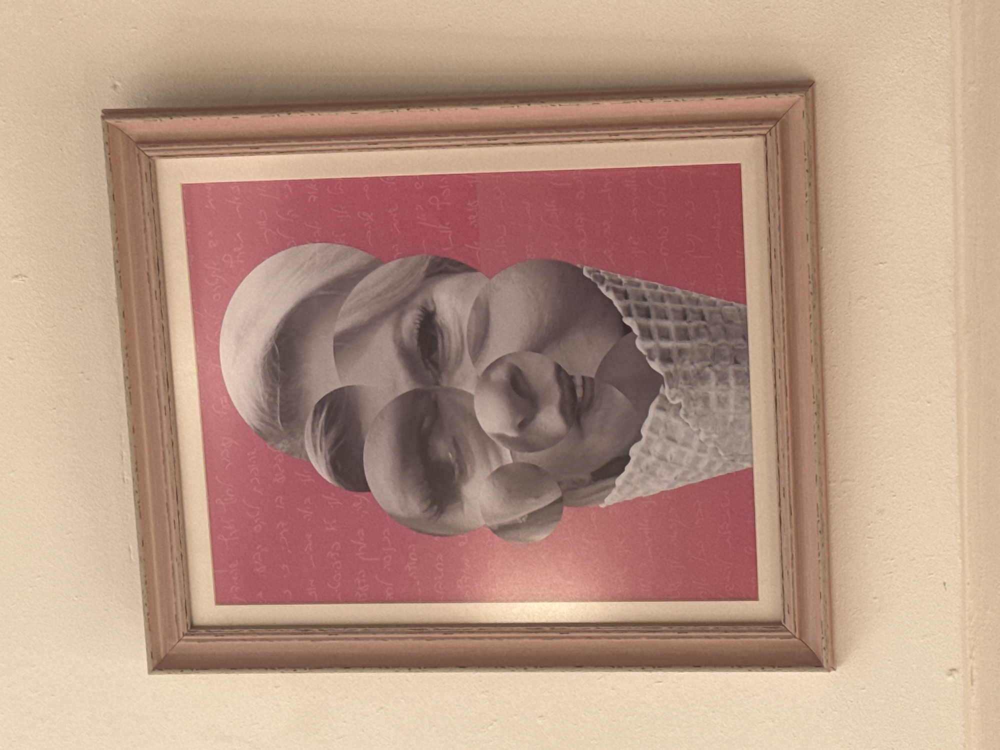 A framed picture hangs on a white wall. The picture features a pink background with faint script. Overlaid on the background is a collage of a woman's face segmented into overlapping circles, one of which transitions into an ice cream cone. The frame is pink with a double-layer design. The location is Tel-Aviv, Israel.