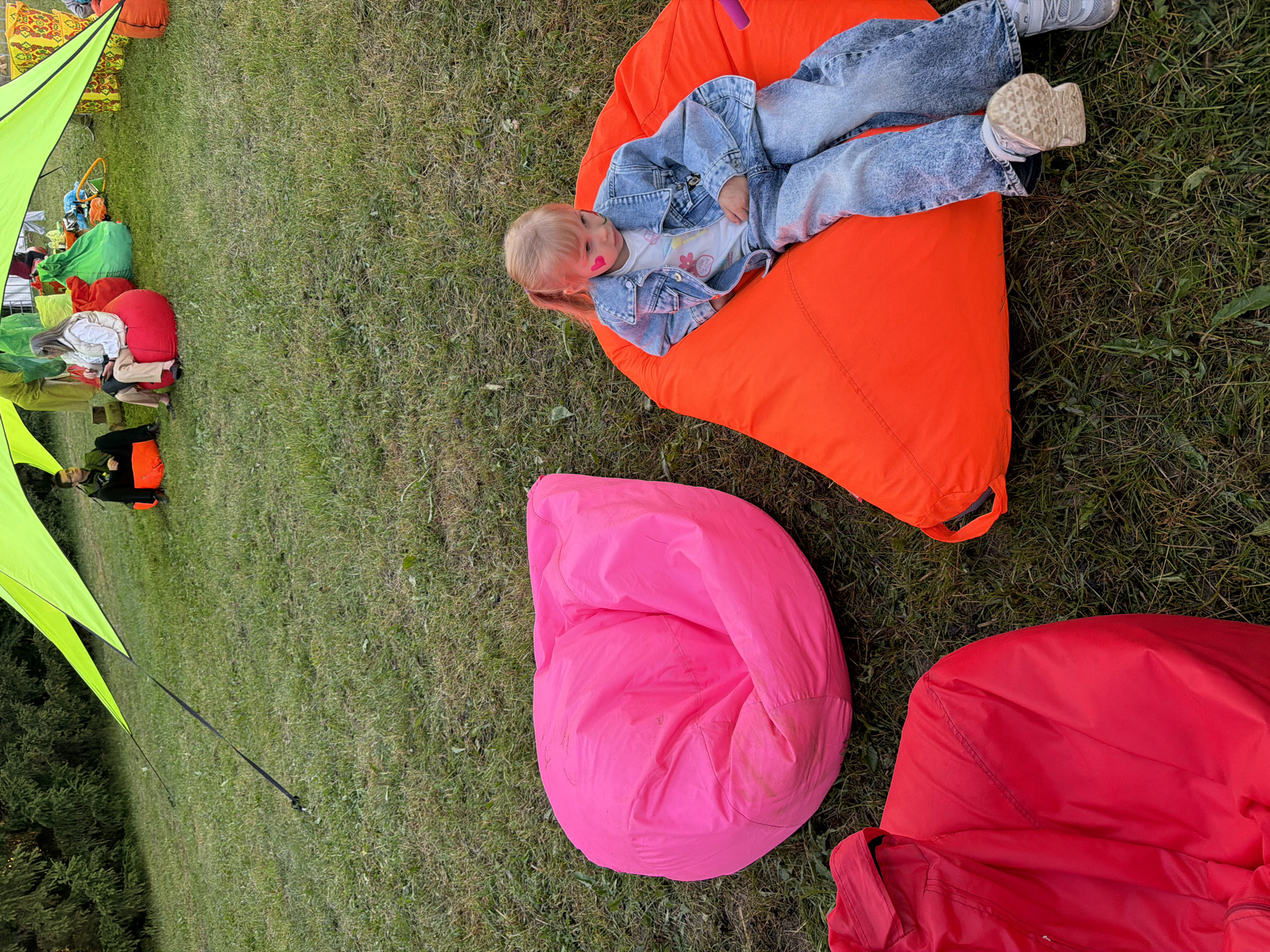 In the city of Unknown, Unknown, a young girl with blonde hair tied in a ponytail, wearing a denim jacket and jeans, sits on a large orange beanbag chair in a grassy outdoor area. A pink heart is visible on her cheek. In the background, several other people are seated on colorful beanbag chairs under a large, bright green canopy. The ground is covered in green grass, and trees are visible in the distance. Two large beanbag chairs, one bright pink and one red, are in the foreground.