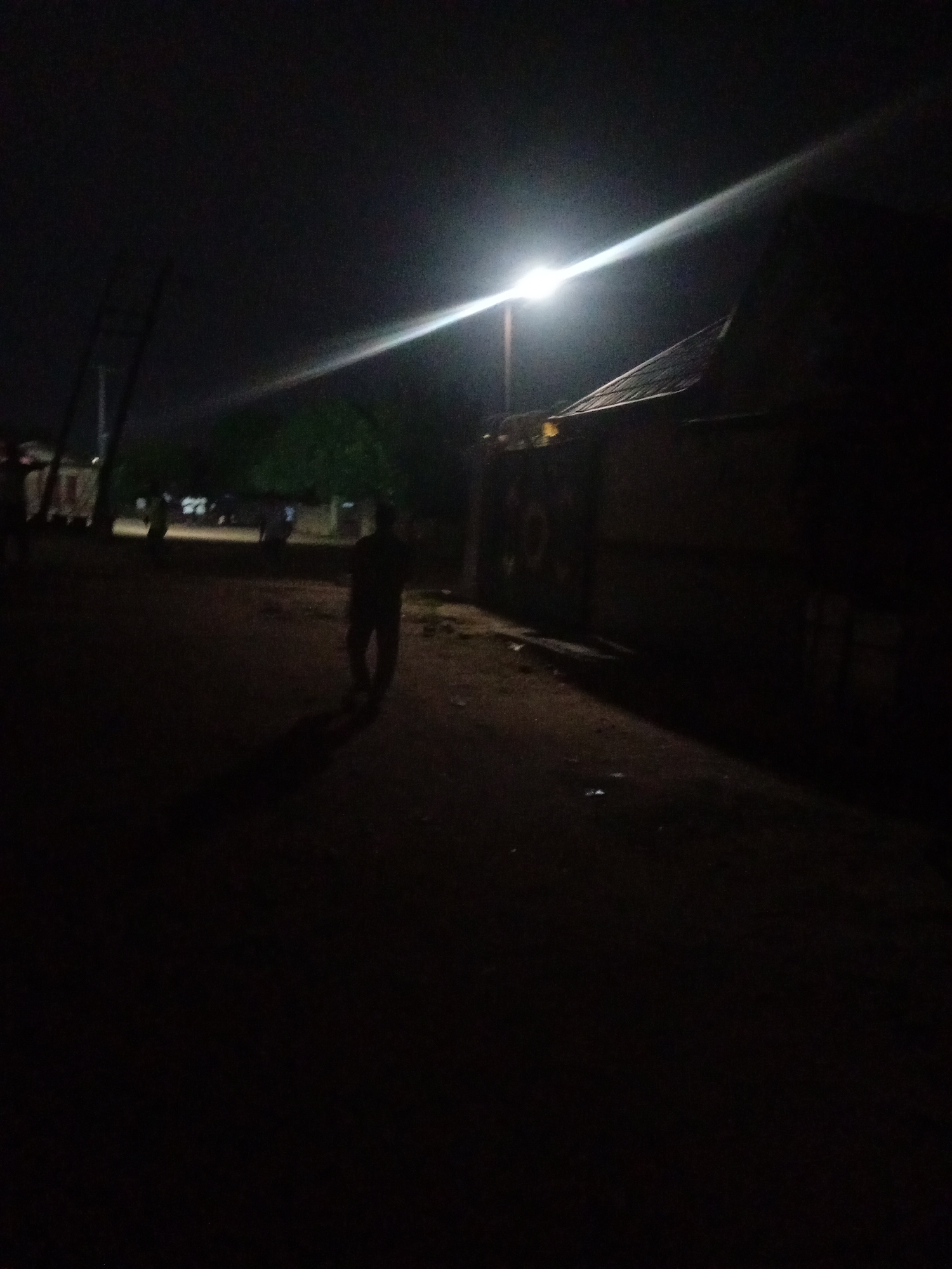The image depicts a nighttime scene in Sabon Gari Nassarawa, Nigeria, illuminated by a bright streetlamp. A lone figure, silhouetted against the light, walks away from the camera on a dirt path. In the background, a modest building with a corrugated roof is visible, partially obscured by darkness. Other indistinct figures and structures can be faintly discerned further in the distance. The overall impression is one of a quiet, rural evening, with the harsh glare of the streetlamp creating strong contrasts between light and shadow. There is no visible text in the image, and no specific activity or event is clearly identifiable beyond the solitary movement of the individual. The time of day is definitively night, and the weather appears to be clear, with no signs of rain or strong wind. The location cues suggest a developing area, possibly a village or the outskirts of a town, given the unpaved road and simple architecture. The emotional tone is ambiguous, leaning towards solitude or perhaps a sense of isolation due to the darkness surrounding the illuminated path.