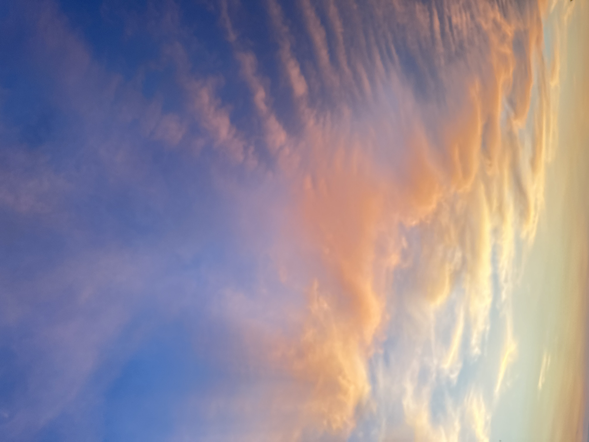 The image shows a vast expanse of sky during what appears to be sunset. Wispy clouds are illuminated with shades of orange, pink, and yellow, transitioning into a deep blue towards the upper portion of the frame. The lower part of the sky displays lighter hues of yellow and pale blue, suggesting the sun is near the horizon. There are no discernible structures, people, or objects in the image, focusing entirely on the atmospheric conditions of the sky.
